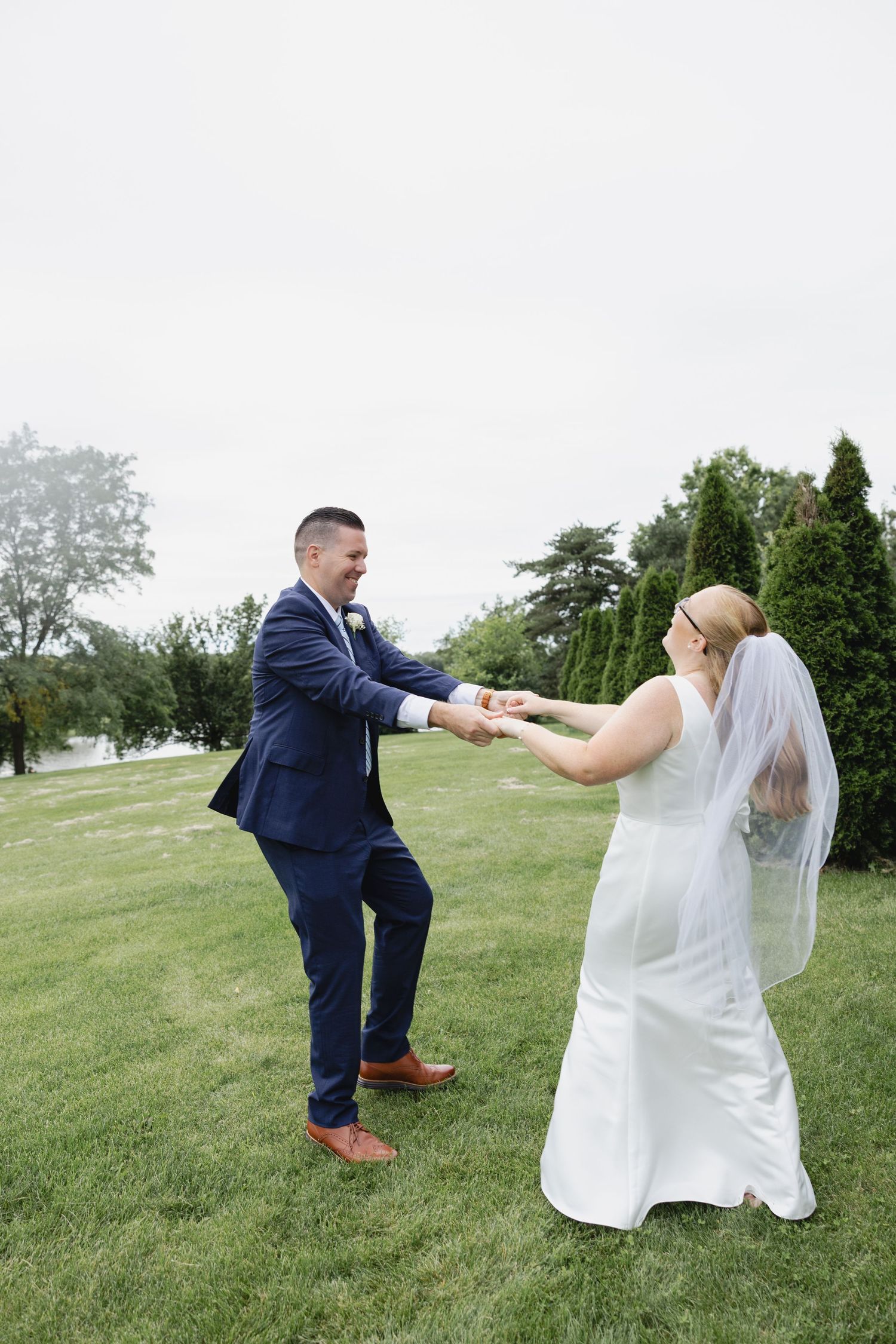 Bride and groom dancing on green lawn, groom in navy suit, bride in white gown with veil, outdoor wedding celebration.