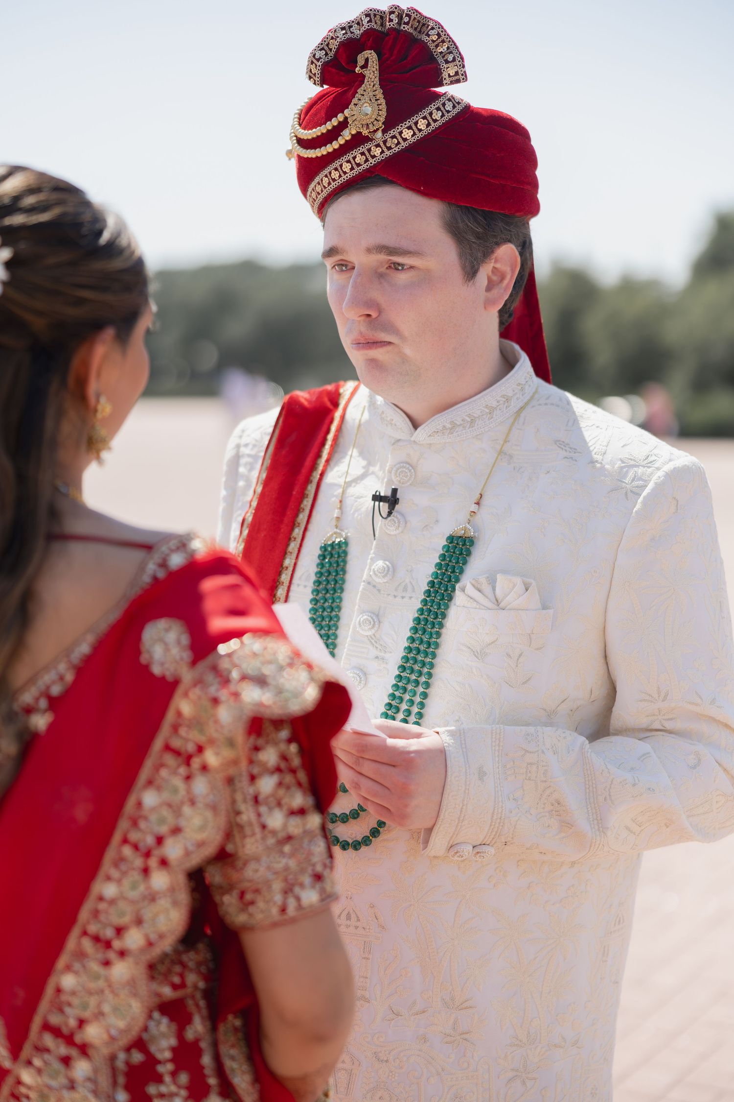 Groom in white sherwani and red turban holding hands with bride in red lehenga at outdoor Indian wedding ceremony.