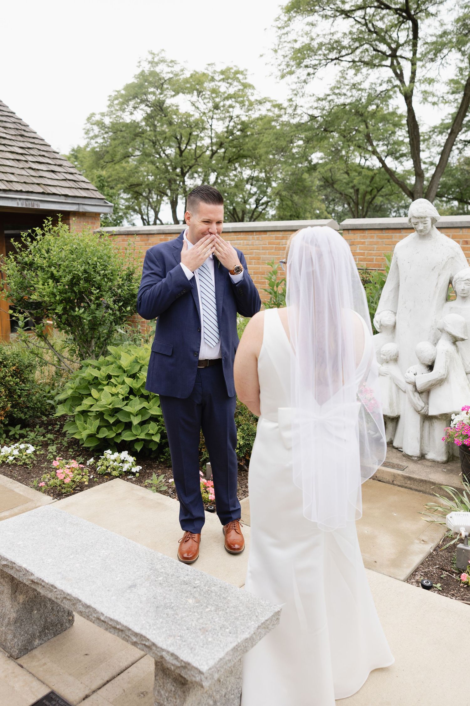 Groom in navy suit reacts emotionally seeing bride in white gown and veil during wedding first look in garden.