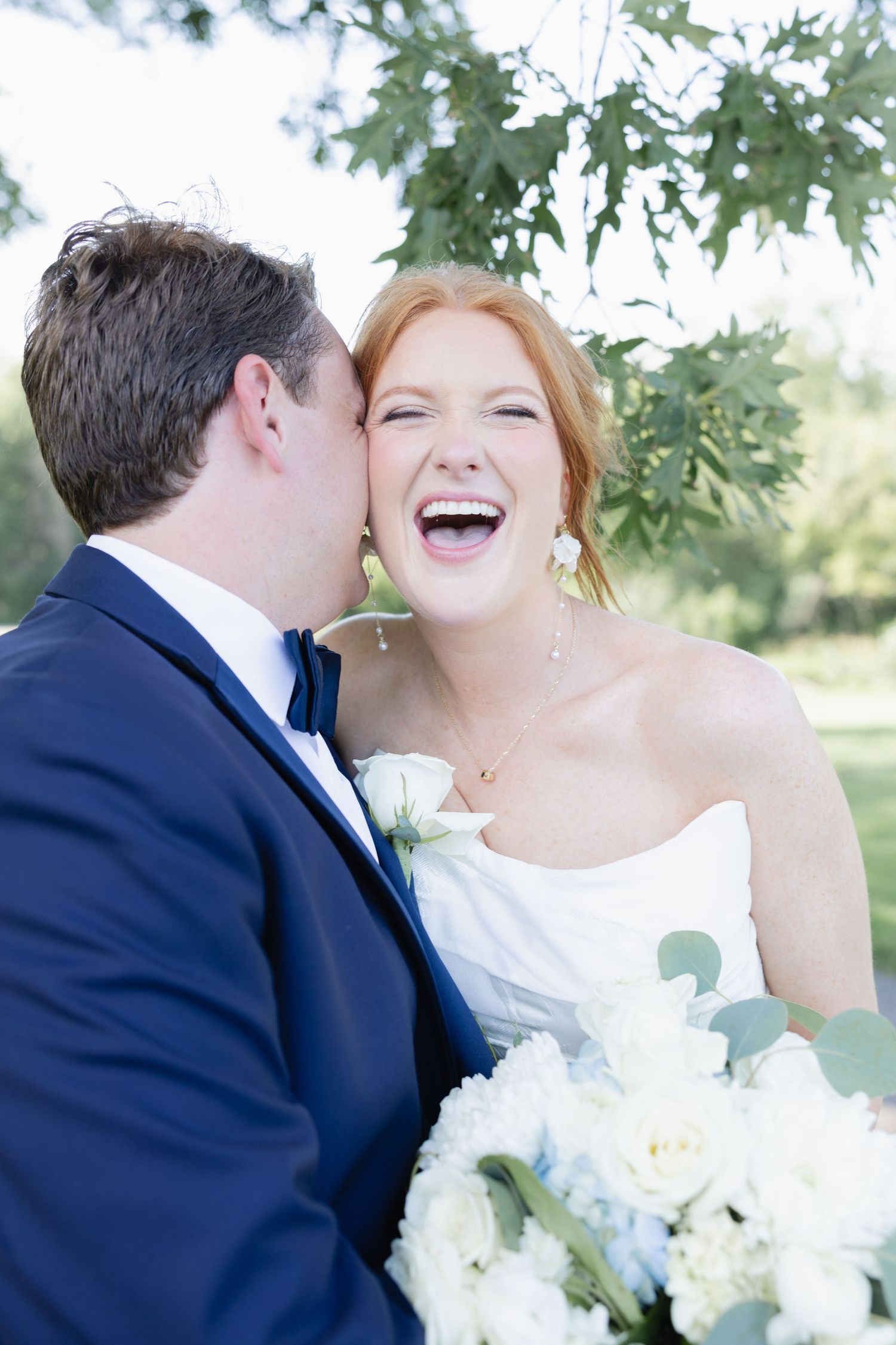 A joyful bride with red hair laughs as her groom in a navy suit kisses her cheek, holding white flowers outdoors.