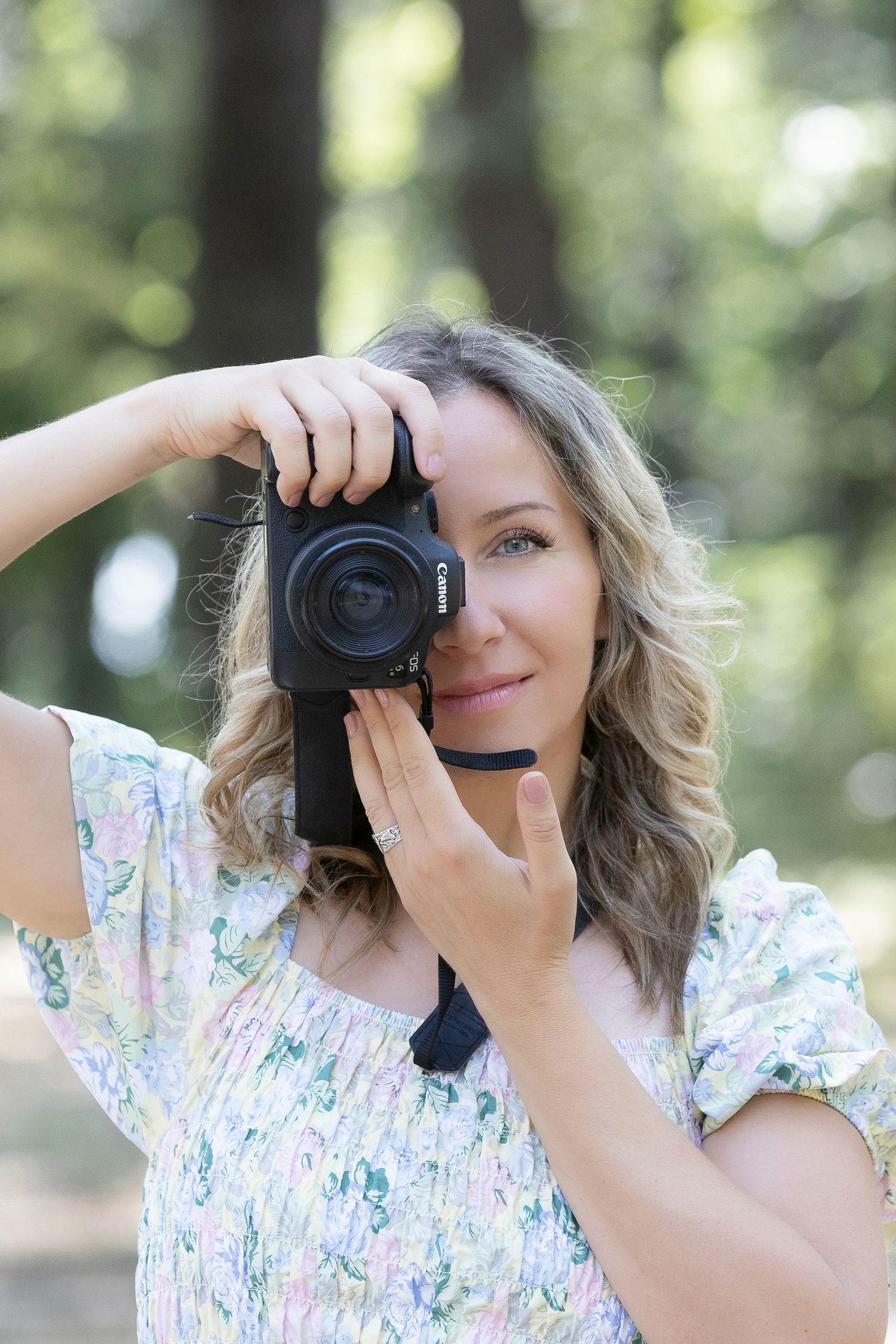 Woman holding a Canon camera up to her face outdoors, wearing a floral dress, with blurred green trees in background.