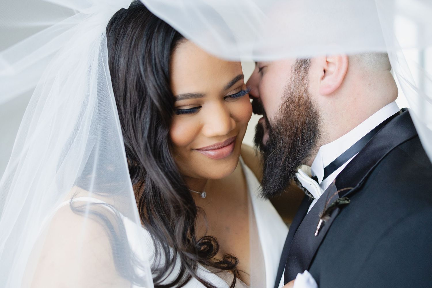 Bride and groom share an intimate moment under a flowing white veil, touching foreheads on their wedding day.