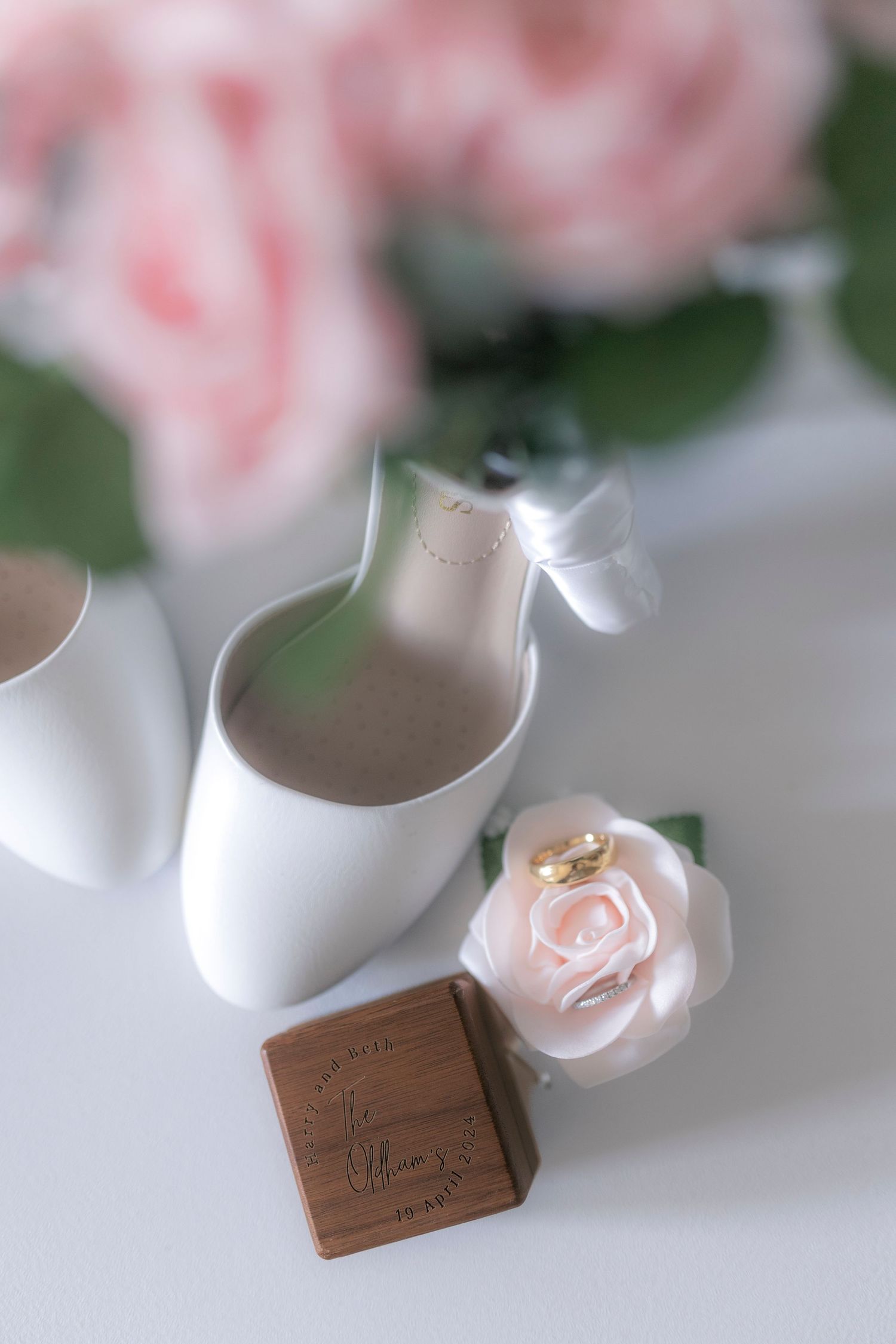 White bridal shoe with pink rose and chocolate wedding favor on white surface.