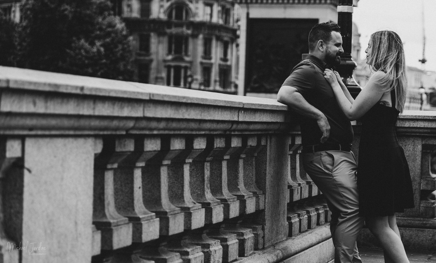 A romantic couple embraces on a balcony with ornate stone railings in this black and white photograph.