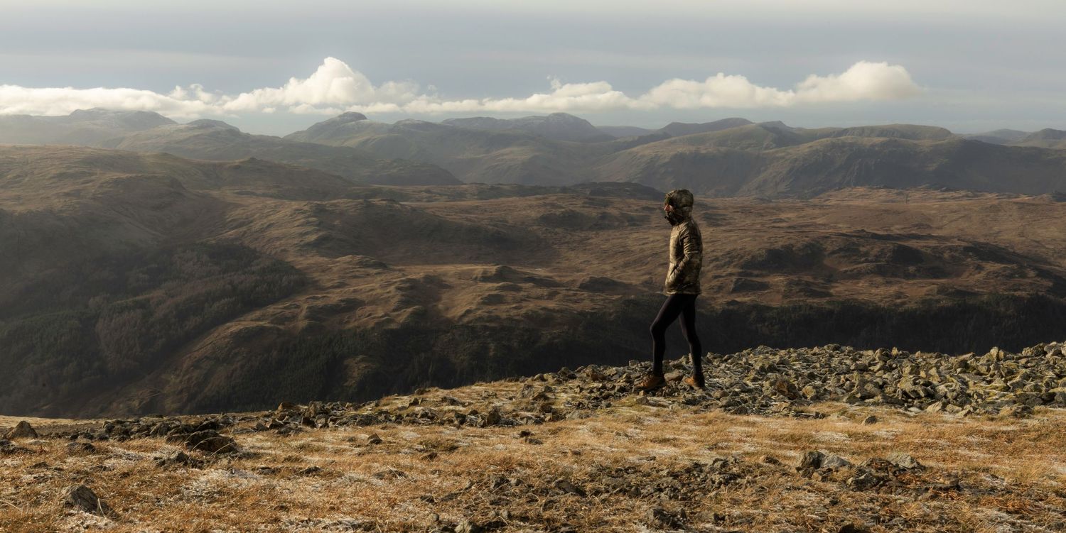 Silhouette of a hiker standing on a rocky mountain summit overlooking vast rolling hills under cloudy skies.