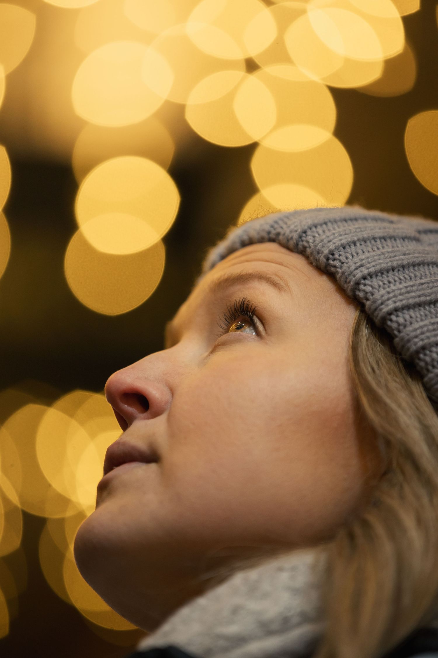 A side profile shows an upward glance against warm bokeh lights while wearing a gray winter hat.