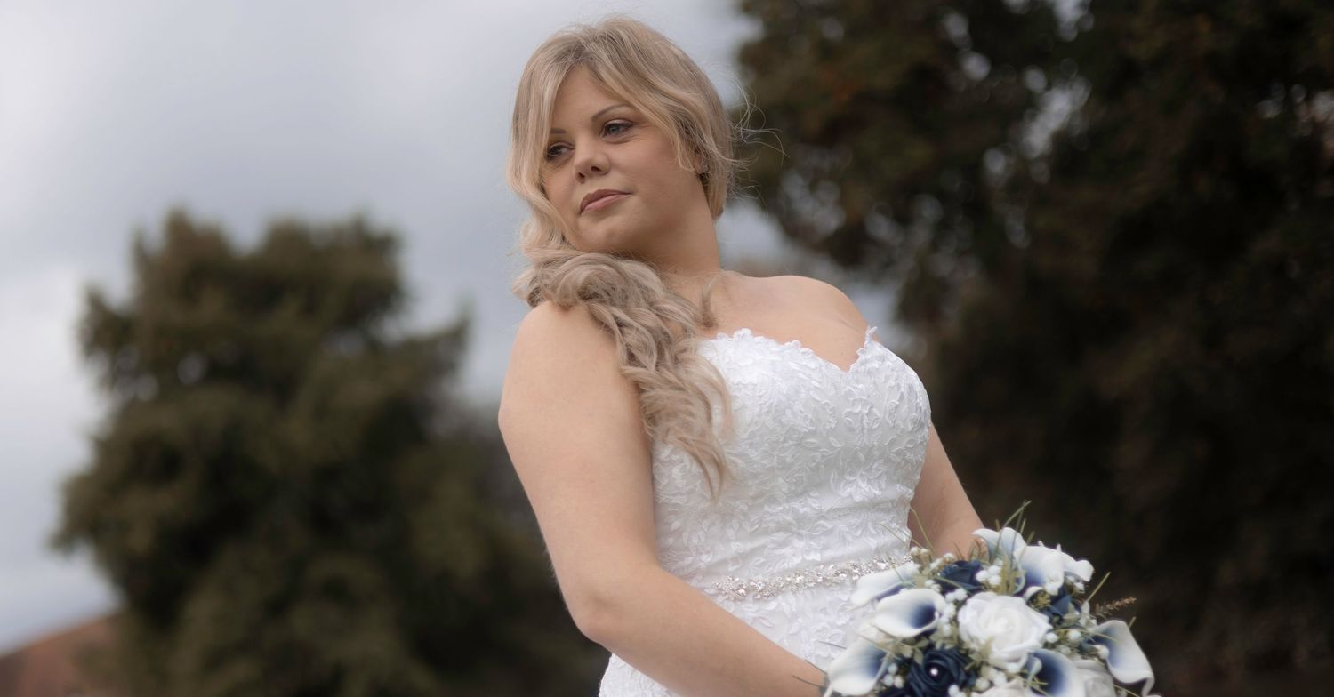 A bride in a white wedding dress holds a bouquet while standing outdoors near trees.
