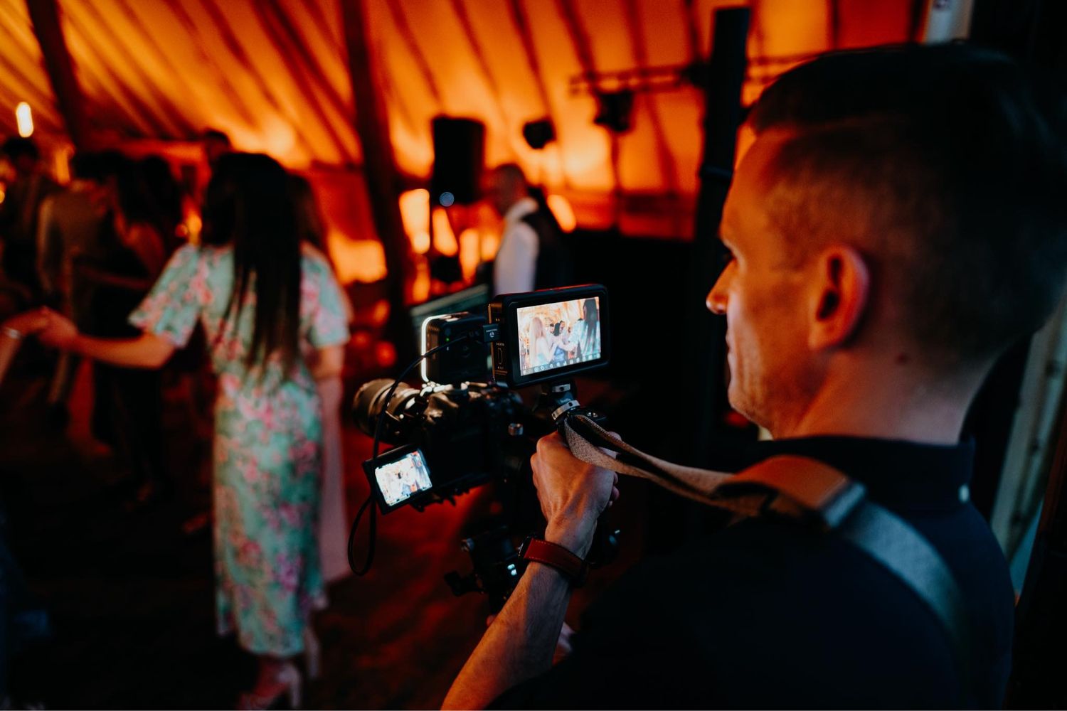 A videographer films a live performance with a professional camera rig in a dimly lit venue with warm lighting.