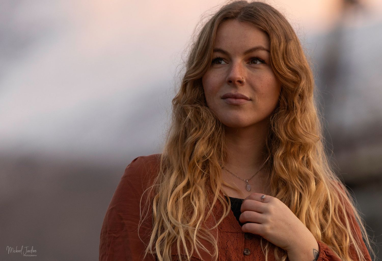 A woman with wavy blonde hair and a rust-colored sweater poses outdoors during golden hour.