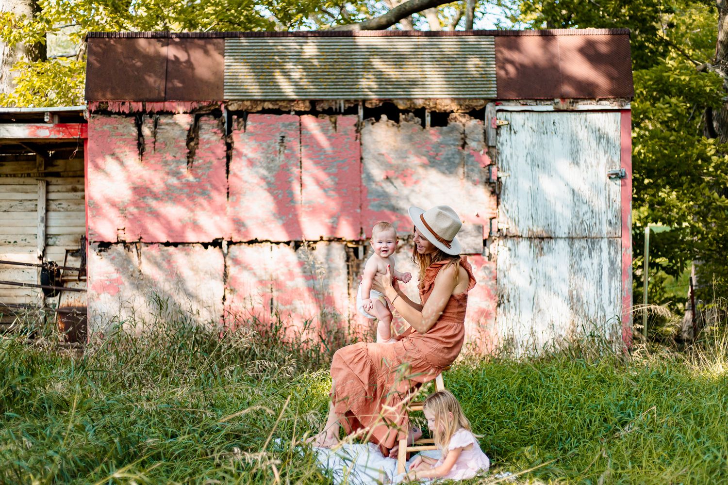 Mom holding her baby son with her toddler daughter at her feet in front of a red run down barn.