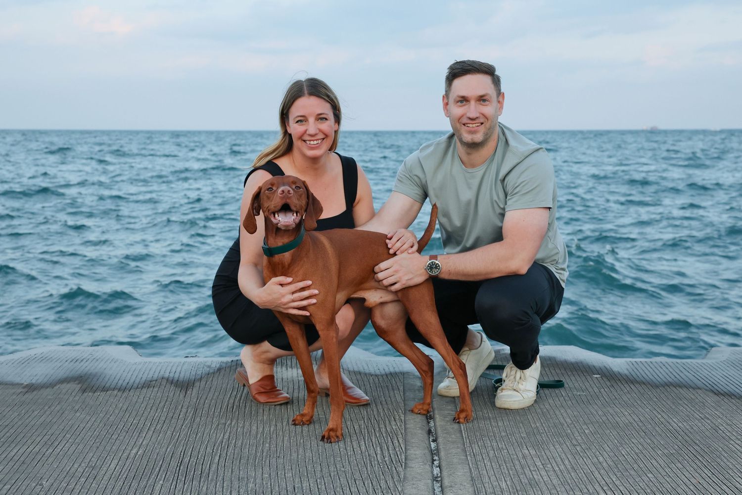 Smiling couple crouching with their Vizsla dog on the lakefront with Lake Michigan in background.
