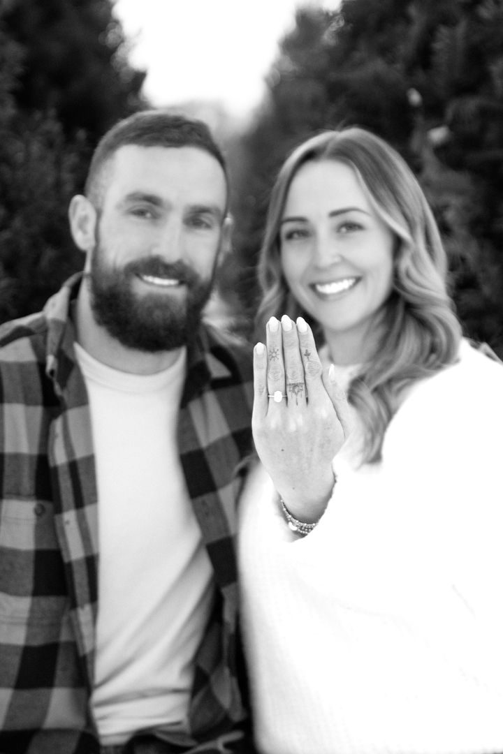 Smiling bearded man in a plaid flannel shirt stands beside a smiling woman showing her engagement ring in a black and white outdoor photo.