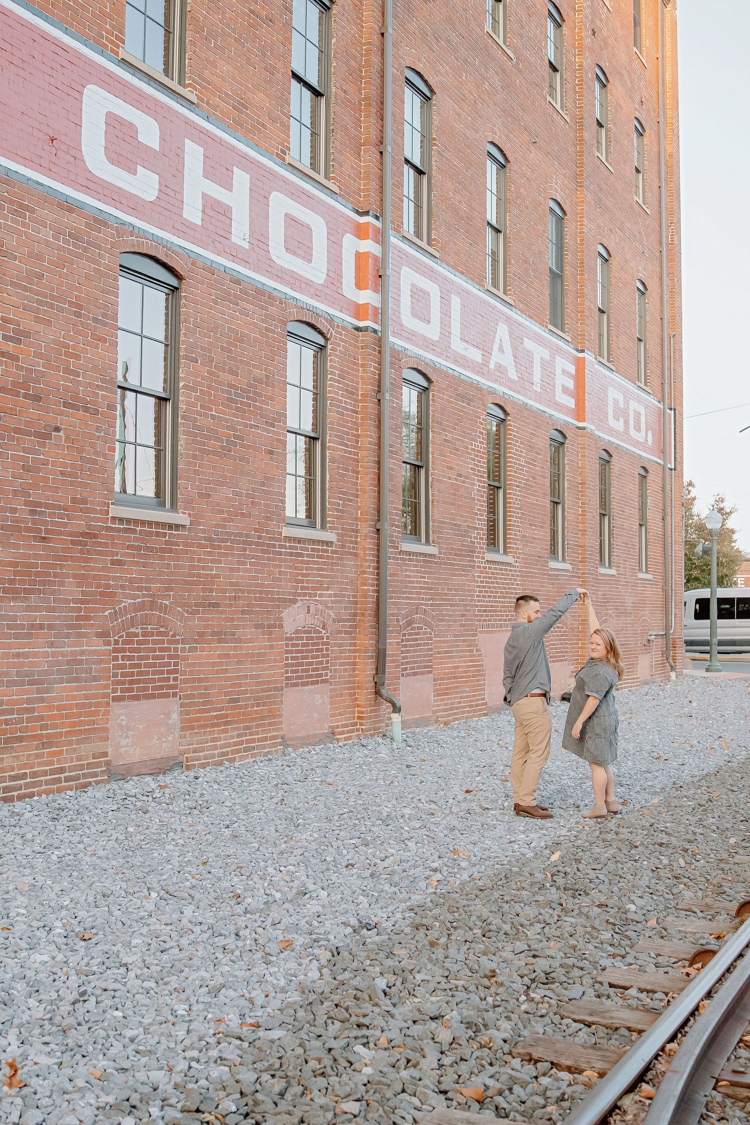 Couple dancing on gravel beside railroad tracks in front of a historic red brick Chocolate Co. building with a faded painted sign.