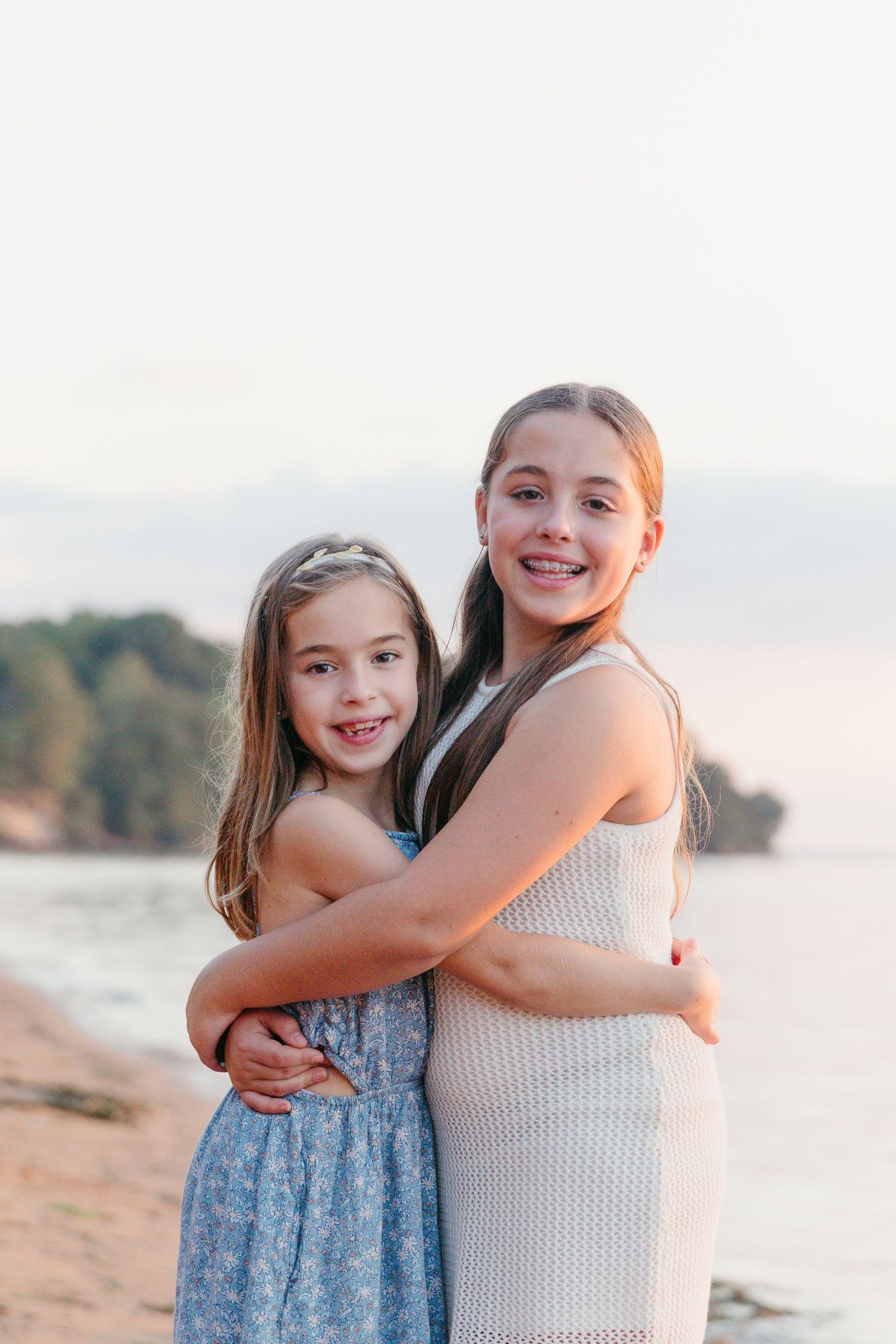 Two smiling girls in summer dresses hug on a sandy beach at sunset, with calm water and tree-covered shoreline behind them.