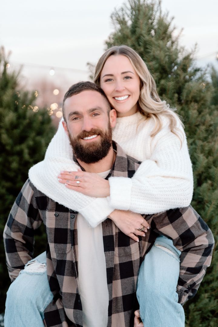Smiling couple in cozy flannel and a white knit sweater embrace in a piggyback pose in front of evergreen trees at a Christmas tree farm.