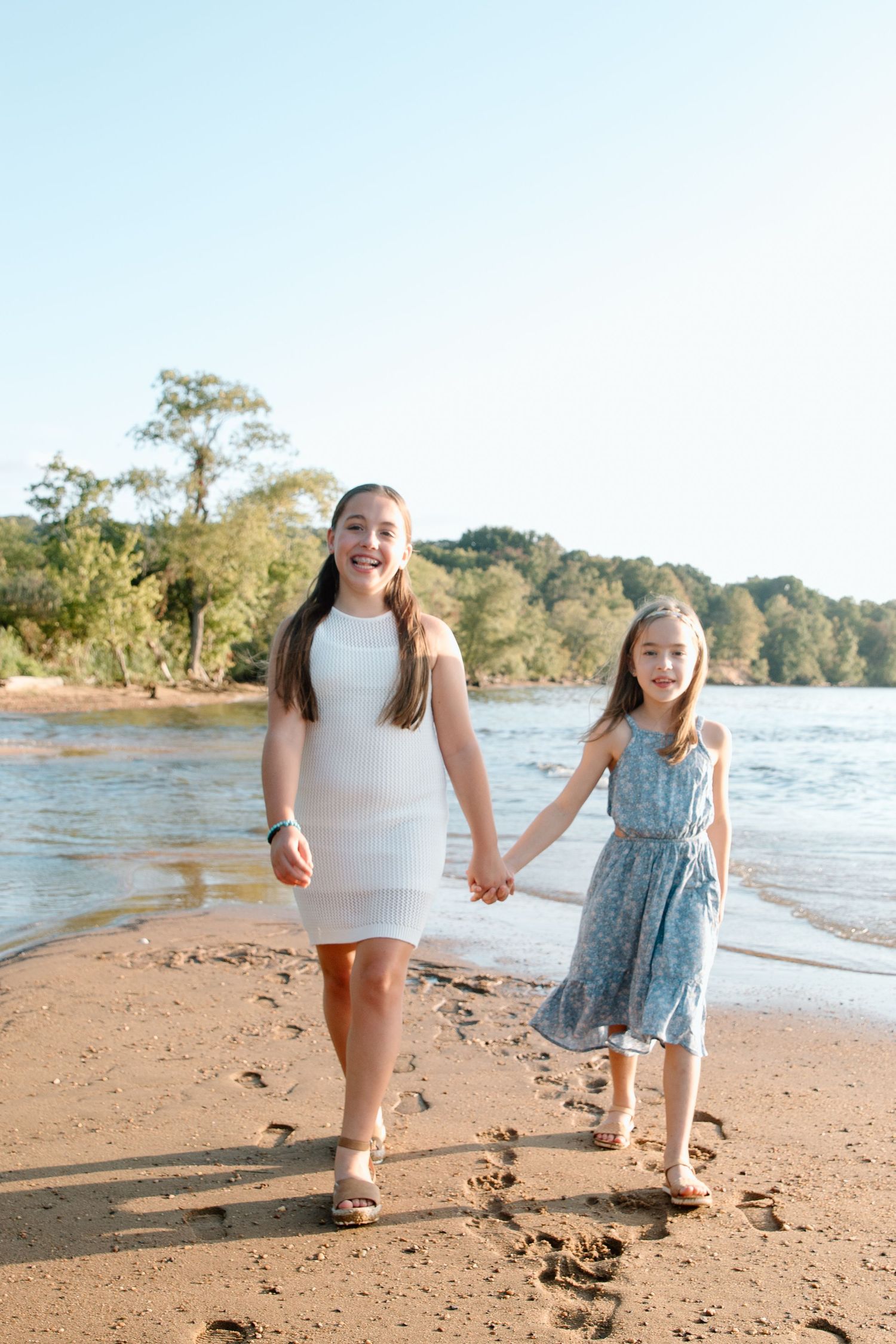 Two smiling girls in summer dresses hold hands while walking along a sandy riverside beach with trees in the background.