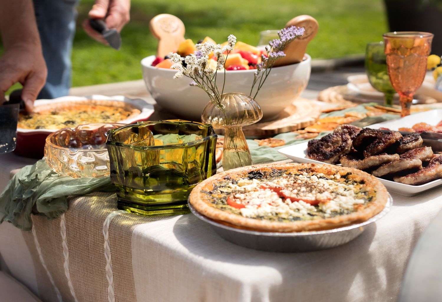 Outdoor picnic table set with a pizza, bread bowl, green glassware, and assorted dishes on a striped linen tablecloth.