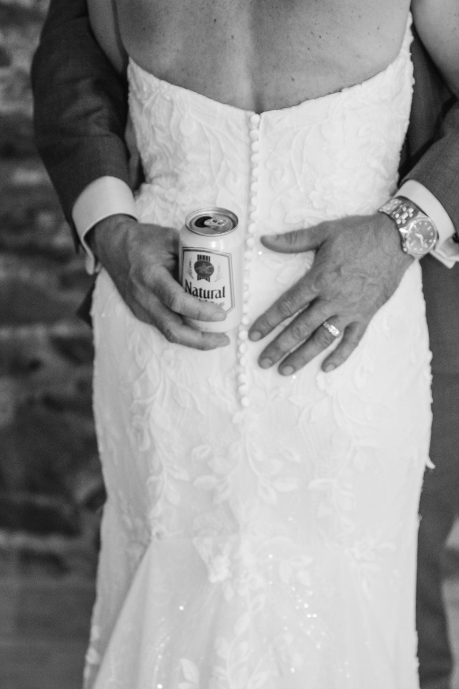 Black and white detail of a groom holding a can of Natural beer while embracing a bride in a button-back lace wedding dress.