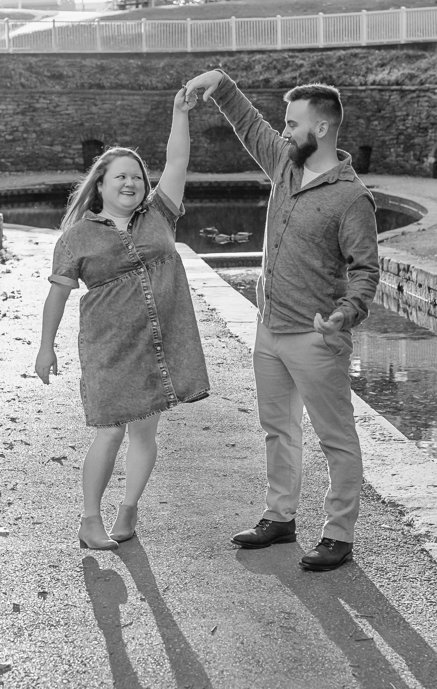 Black and white photo of a man twirling a smiling woman in a denim dress beside a pond during an engagement session.