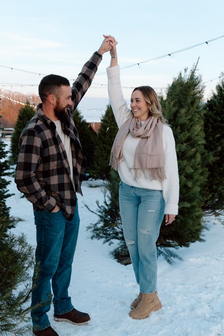 Smiling couple in cozy winter outfits dance hand in hand on snow among evergreen trees at a Christmas tree farm under string lights.