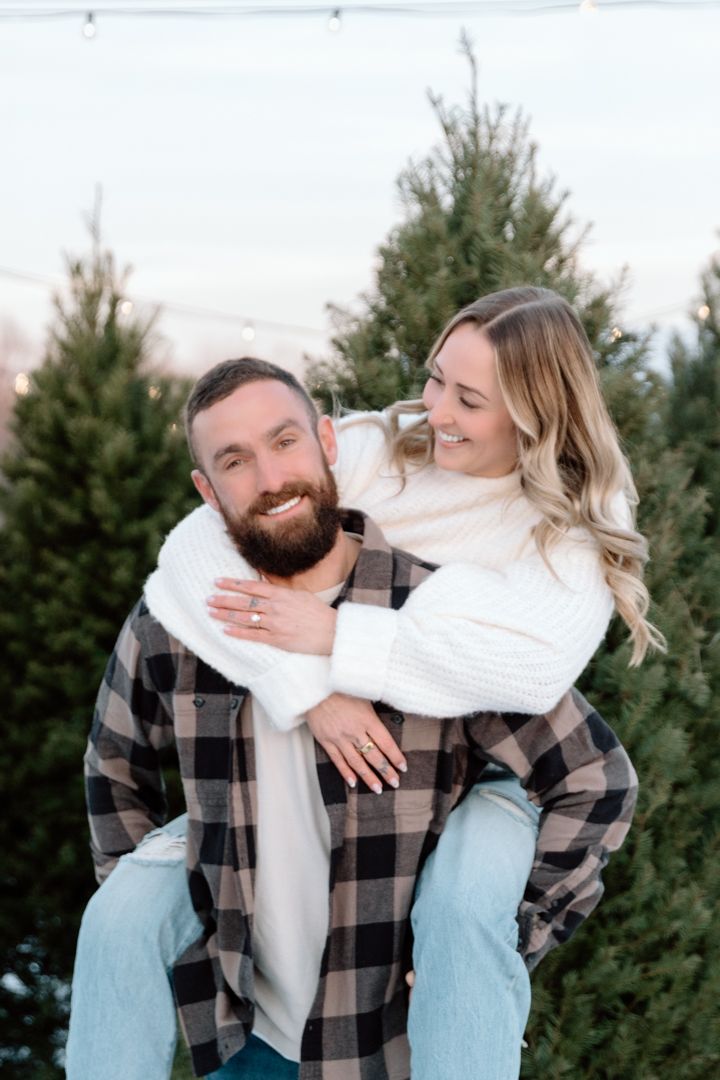 Bearded man gives a laughing woman a piggyback ride among evergreen Christmas trees and string lights, showing her engagement ring.