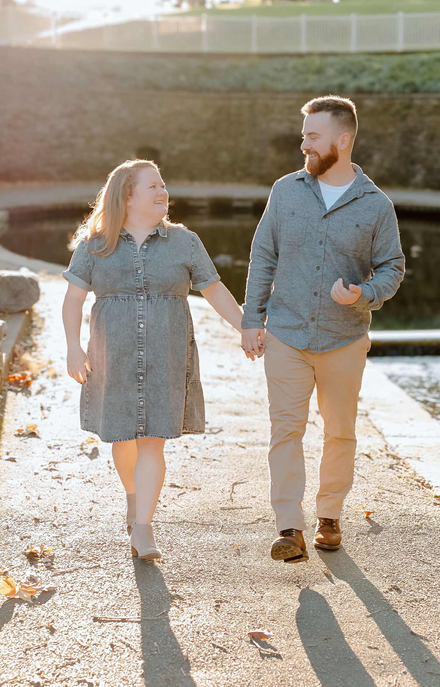 Smiling couple holding hands and walking along a sunlit paved path beside a pond, wearing casual denim and tan outfits on an autumn day.