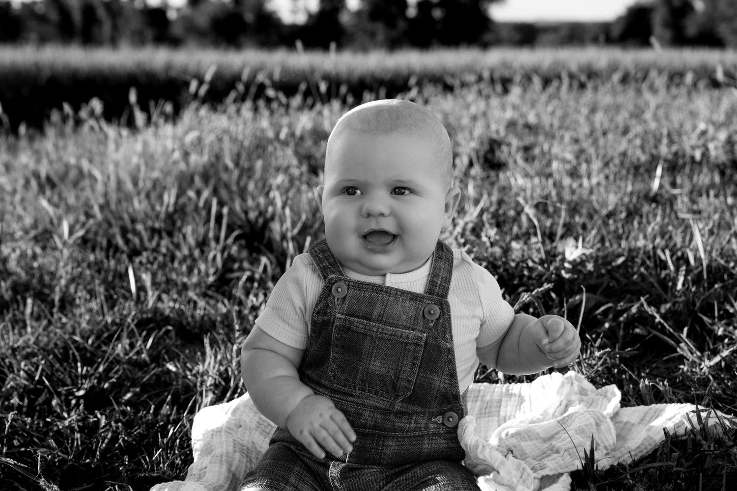 Black and white photo of a smiling baby in denim overalls sitting on a blanket in a grassy field outdoors.
