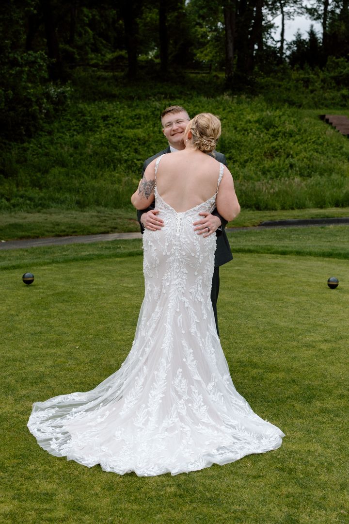 Bride in a lace wedding gown with a low open back embraces a smiling groom in a dark suit on a green lawn at an outdoor wedding.