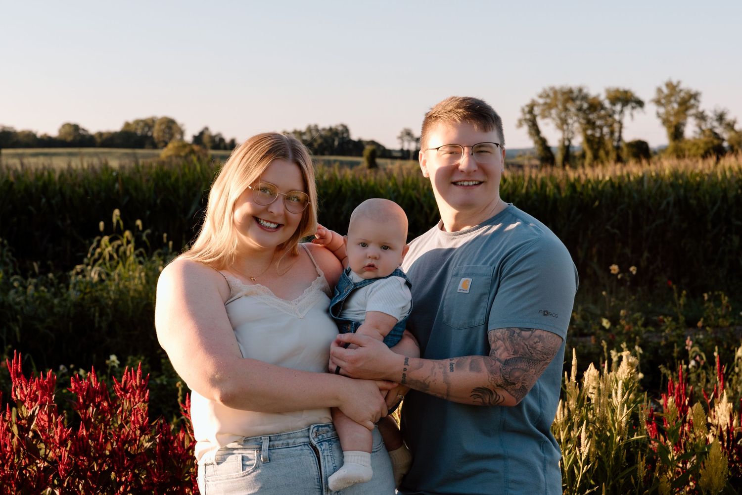 Smiling couple holding their baby while standing in a sunny field with green crops and red flowers at golden hour.