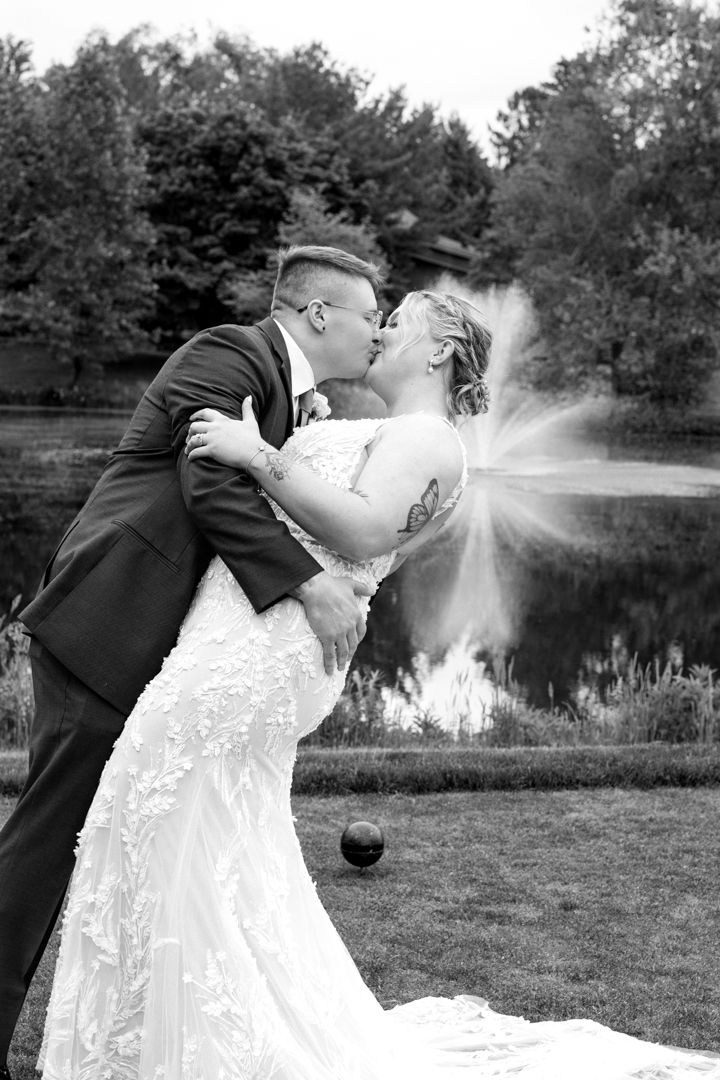 Black and white photo of a groom dipping his bride for a kiss on a lawn beside a pond with a fountain behind them.