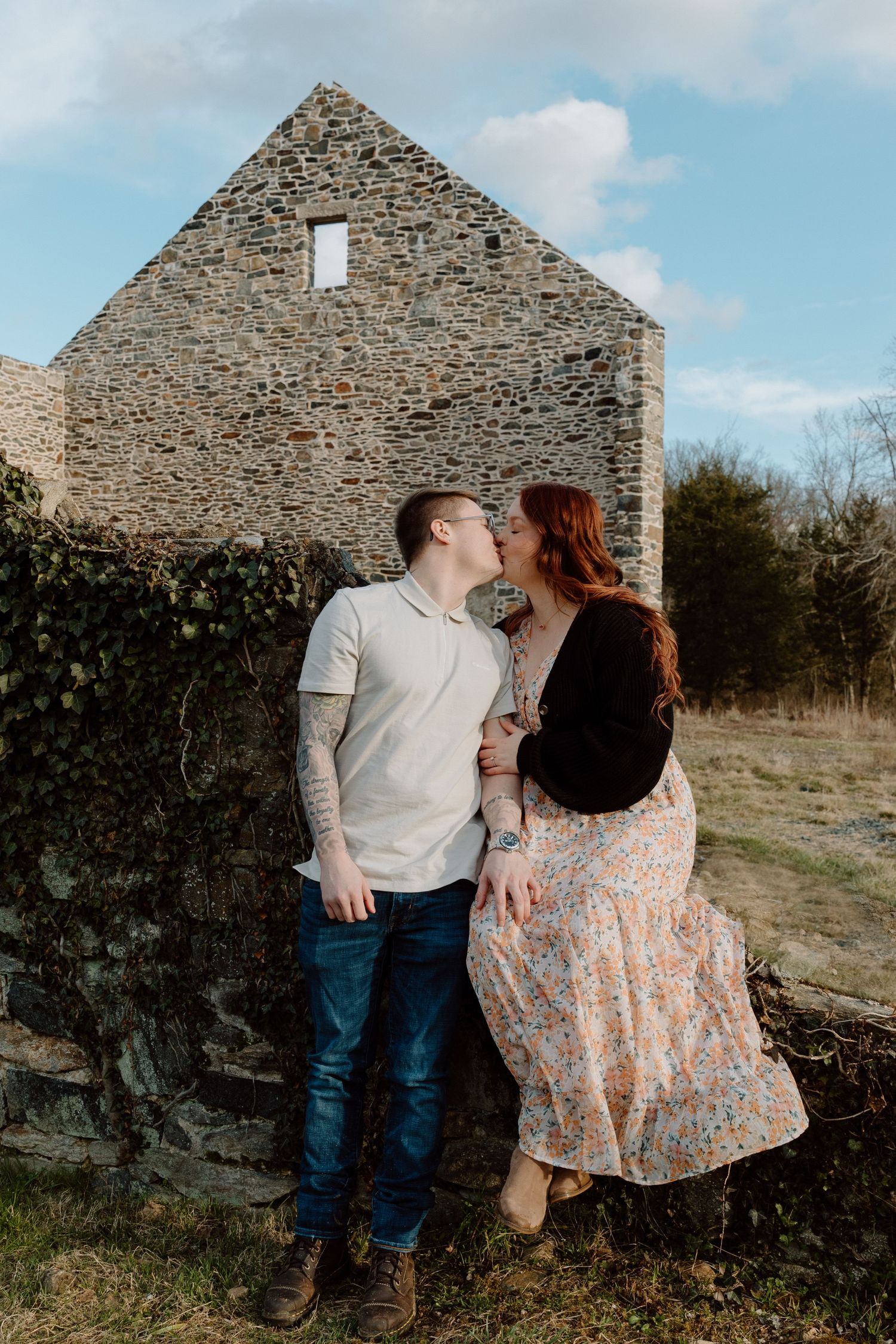 Couple sharing a kiss while sitting on an ivy-covered stone wall in front of a rustic stone building at sunset.