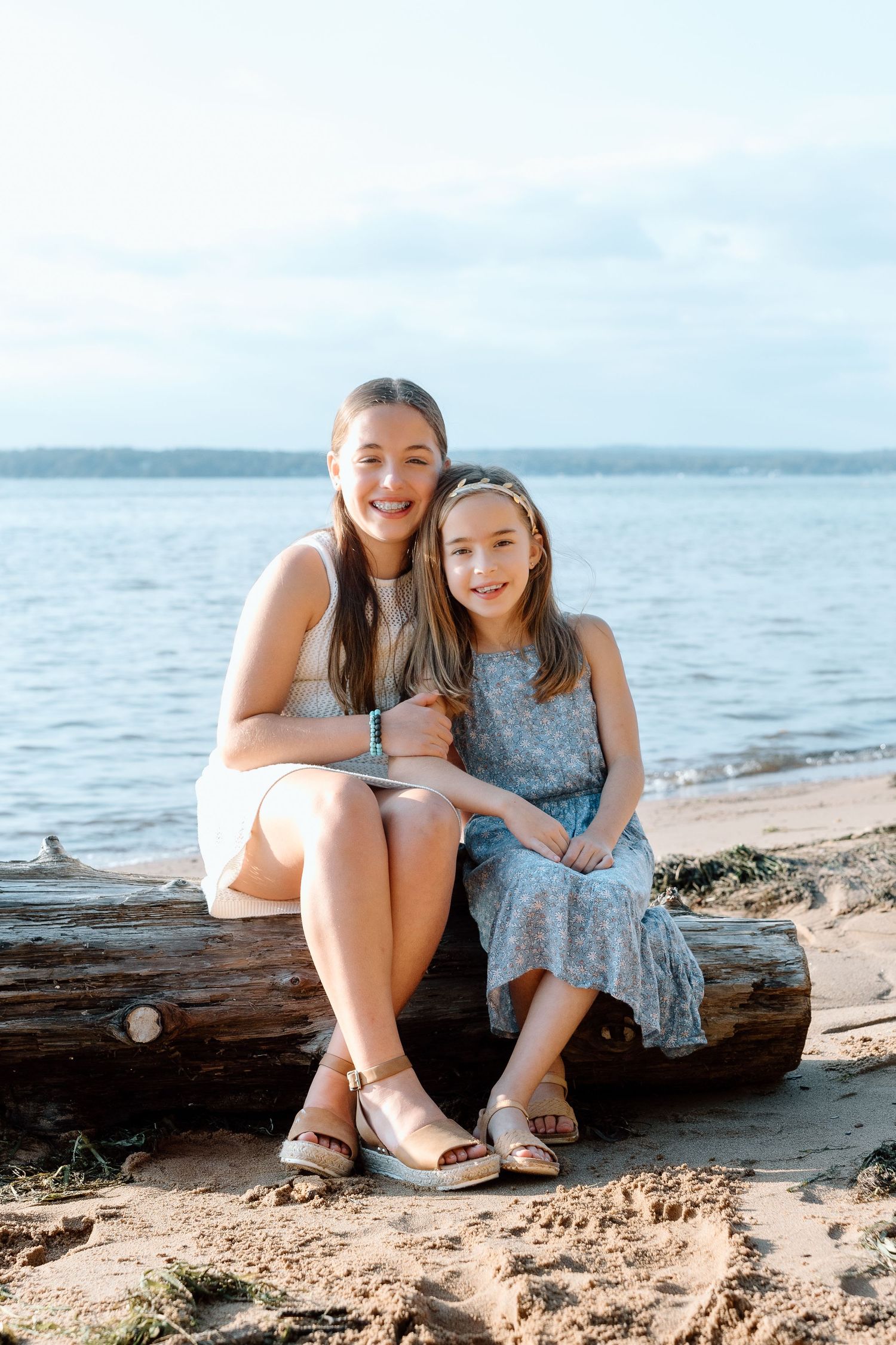 Two smiling girls sit close together on a weathered driftwood log on a sandy beach, with calm water and a soft blue sky behind them.