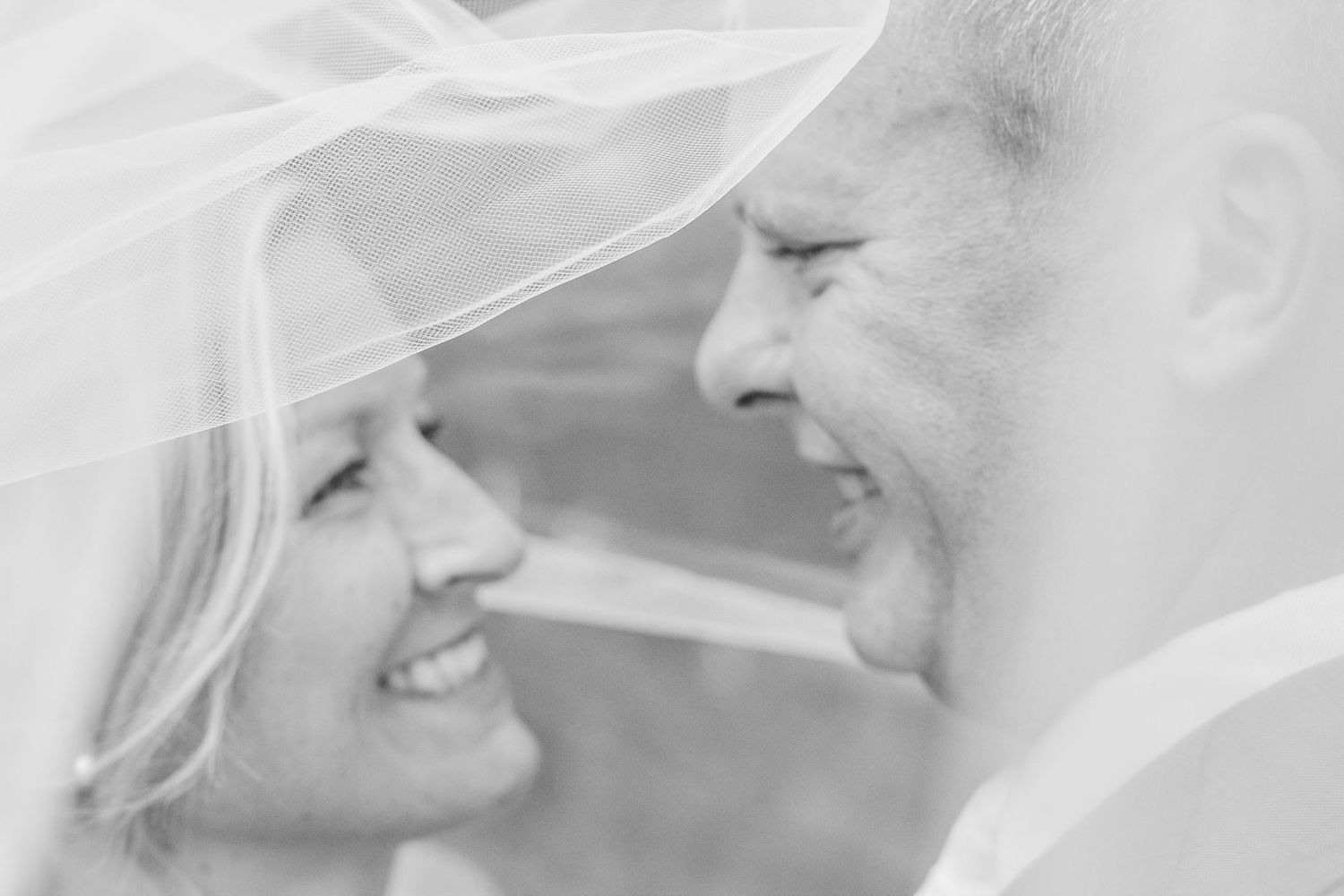 Smiling bride and groom gaze at each other under a sheer wedding veil in a joyful black and white close-up portrait.