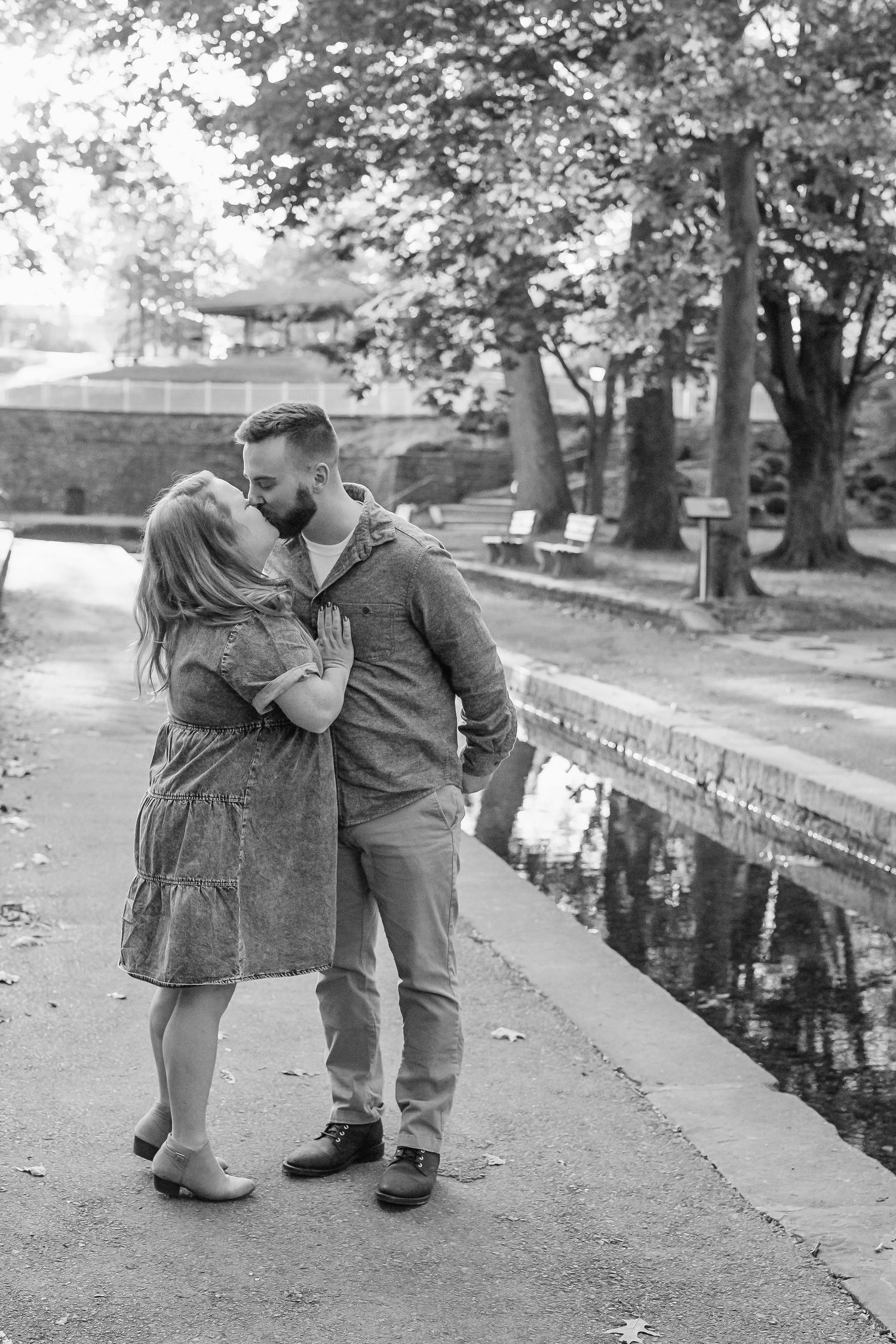 Black and white photo of a couple kissing on a park path beside a reflective canal, with trees and benches in the background.