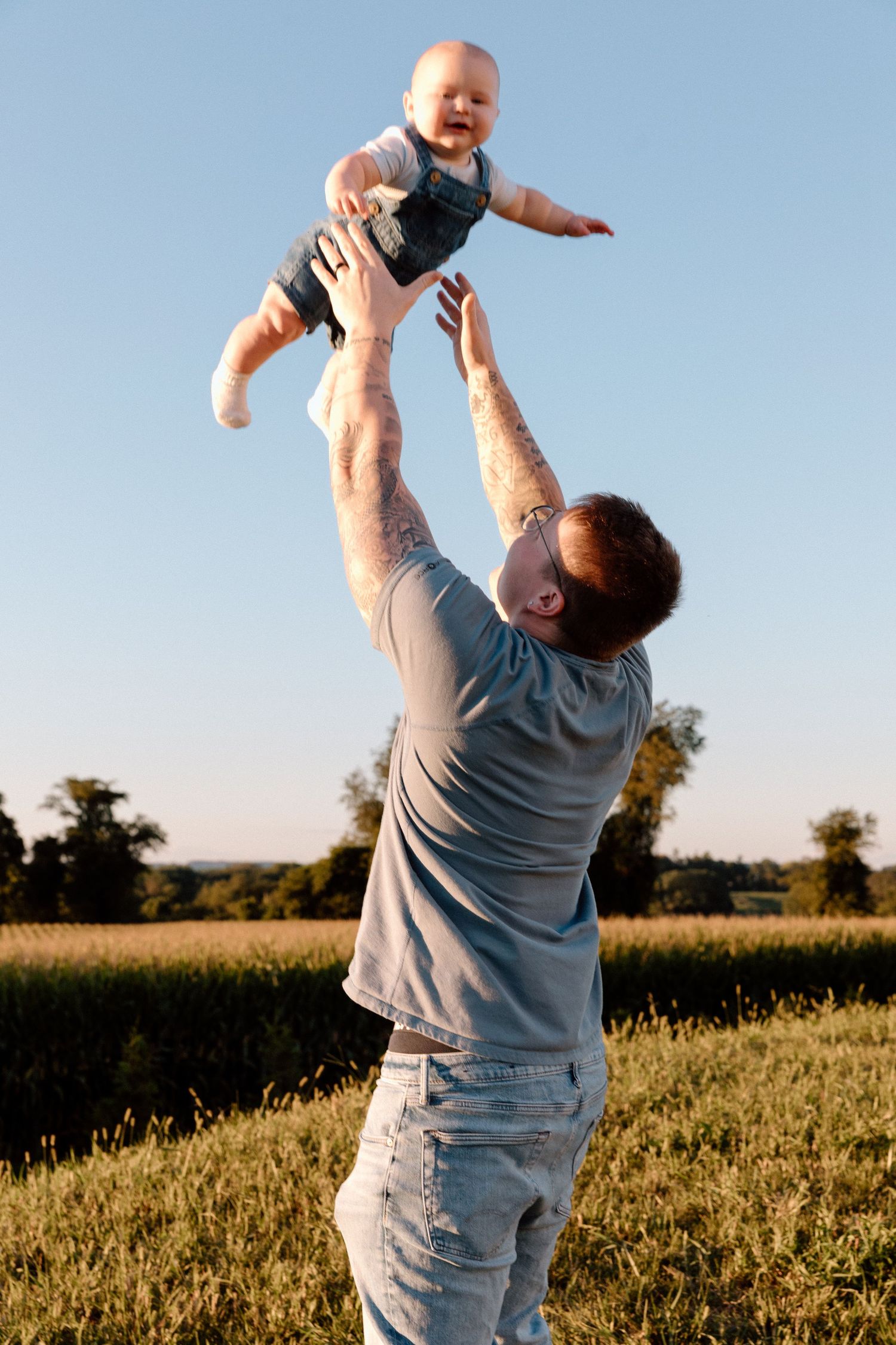 Tattooed father lifts his smiling baby into the air in a sunny grassy field with trees and a clear blue sky.