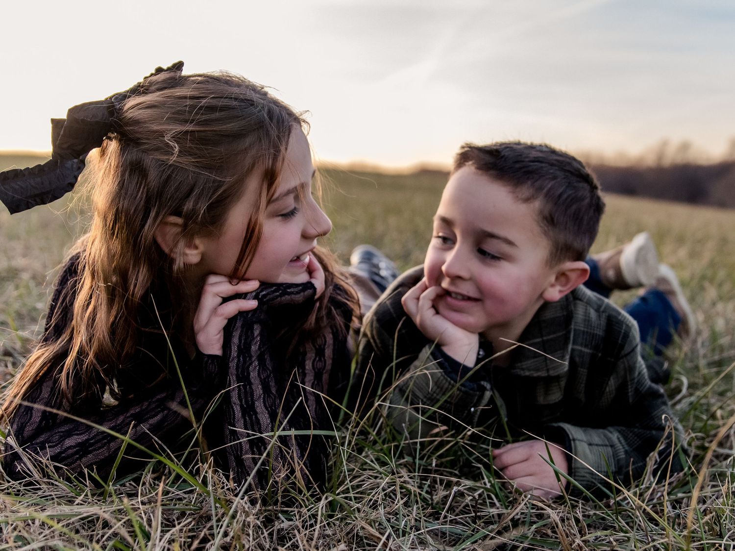 Two smiling children lie on their stomachs in a grassy field at sunset, chins resting on their hands as they chat face to face.