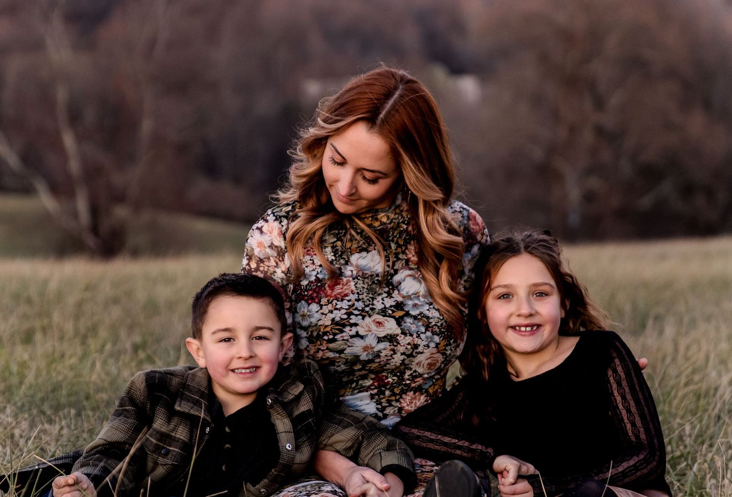 A smiling woman in a floral top sits in a grassy field with a young boy and a smiling girl during a golden-hour family photo session.