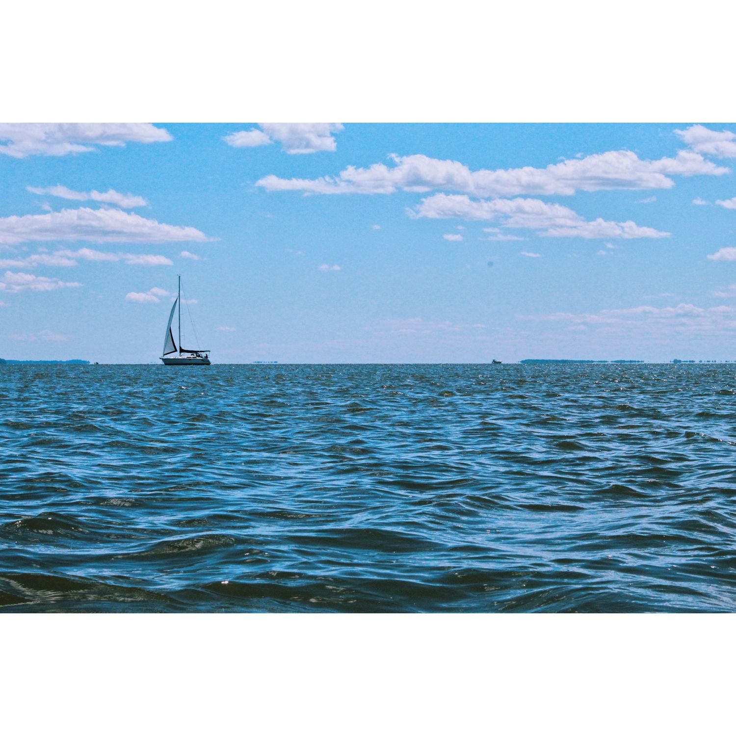 A lone sailboat glides across the choppy blue waters of a wide bay under a bright sky dotted with fluffy white clouds.