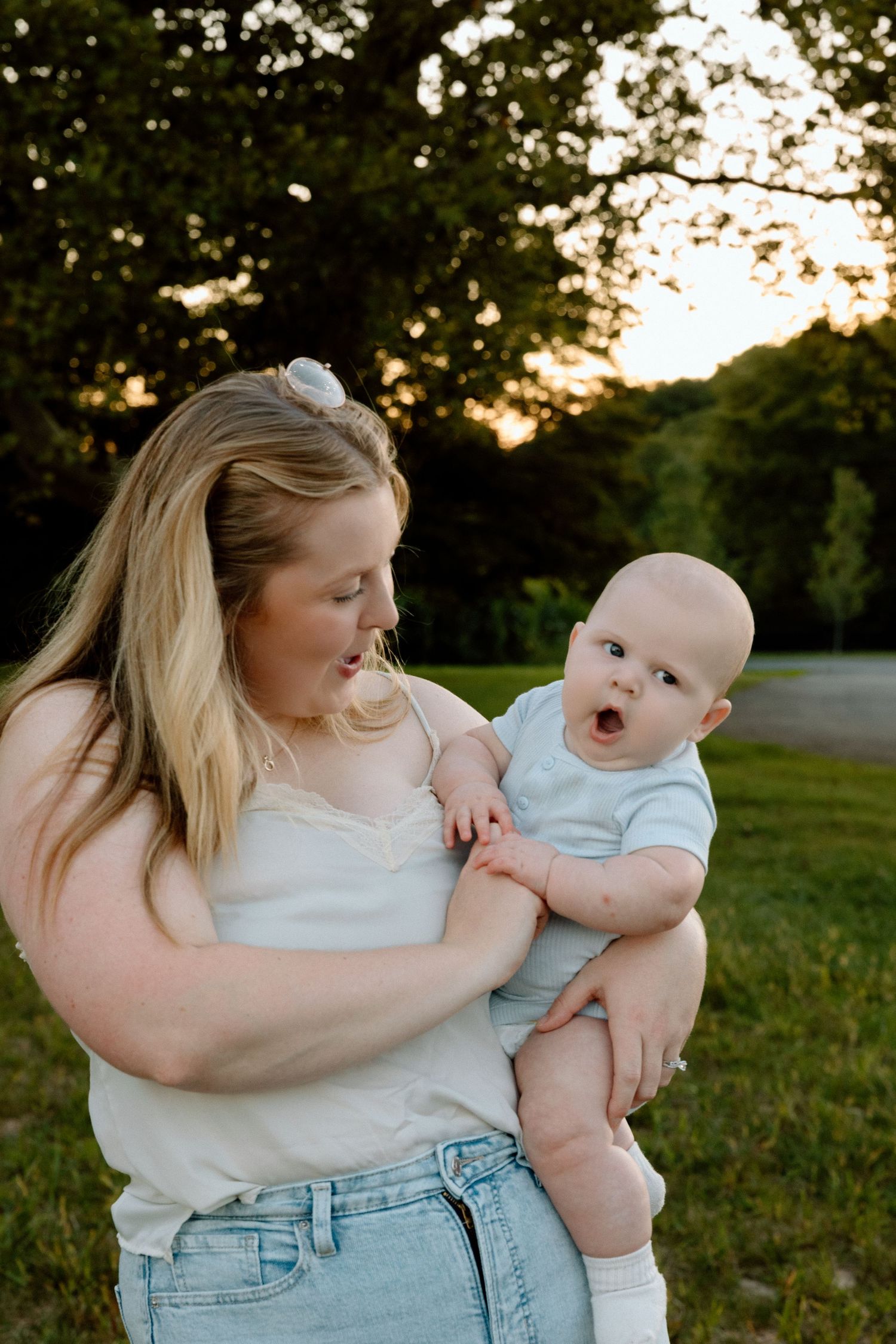 A smiling woman with long blonde hair holds a wide-eyed baby in a light outfit on a grassy lawn at sunset, framed by green trees.