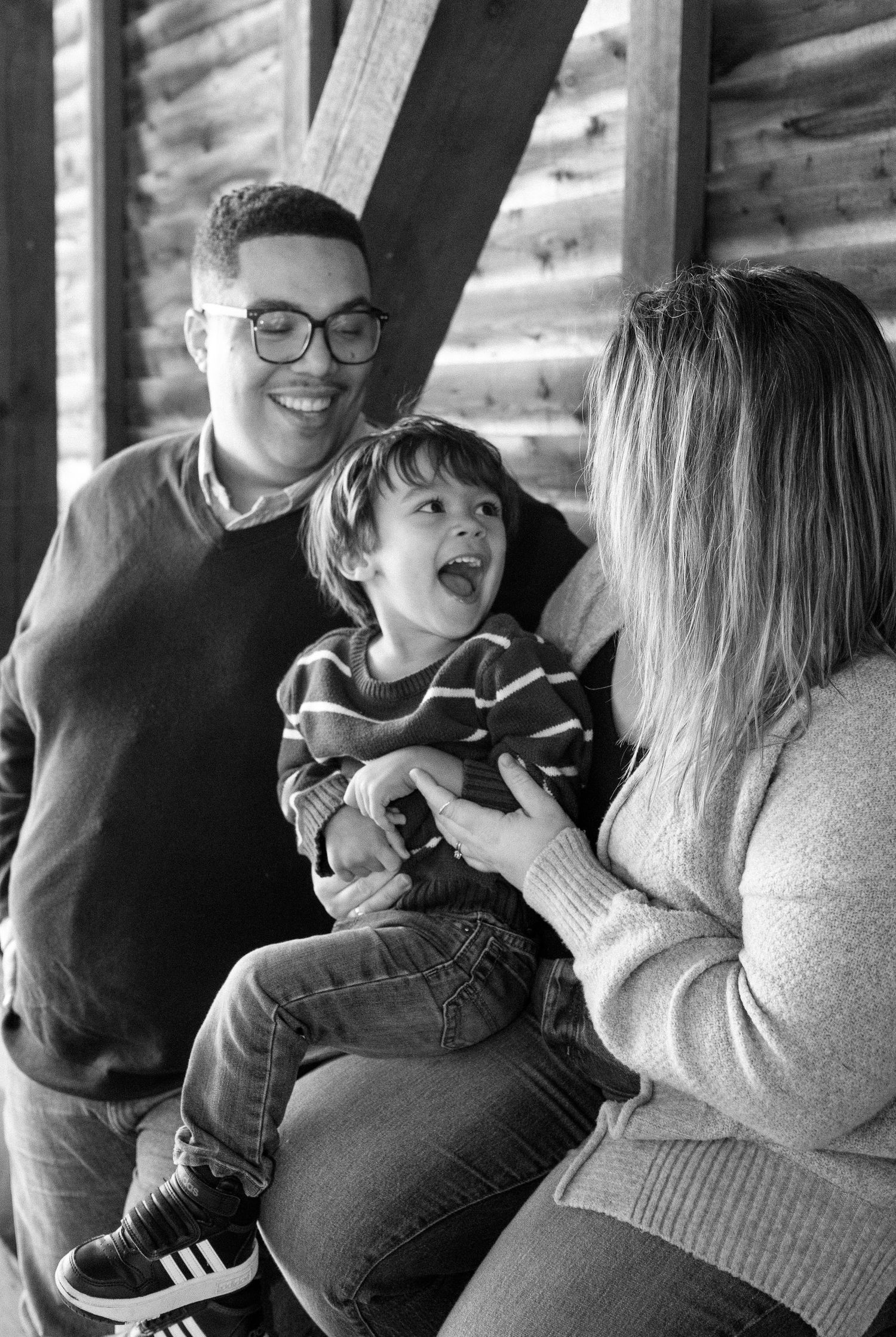 Black and white family portrait of a smiling man with glasses, a woman, and a laughing young boy sitting on his lap by a rustic wooden wall.