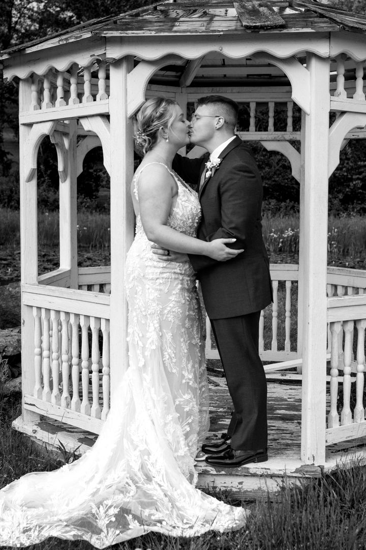A bride in a lace gown and a groom in a suit kiss in a white gazebo in this black and white wedding photo.