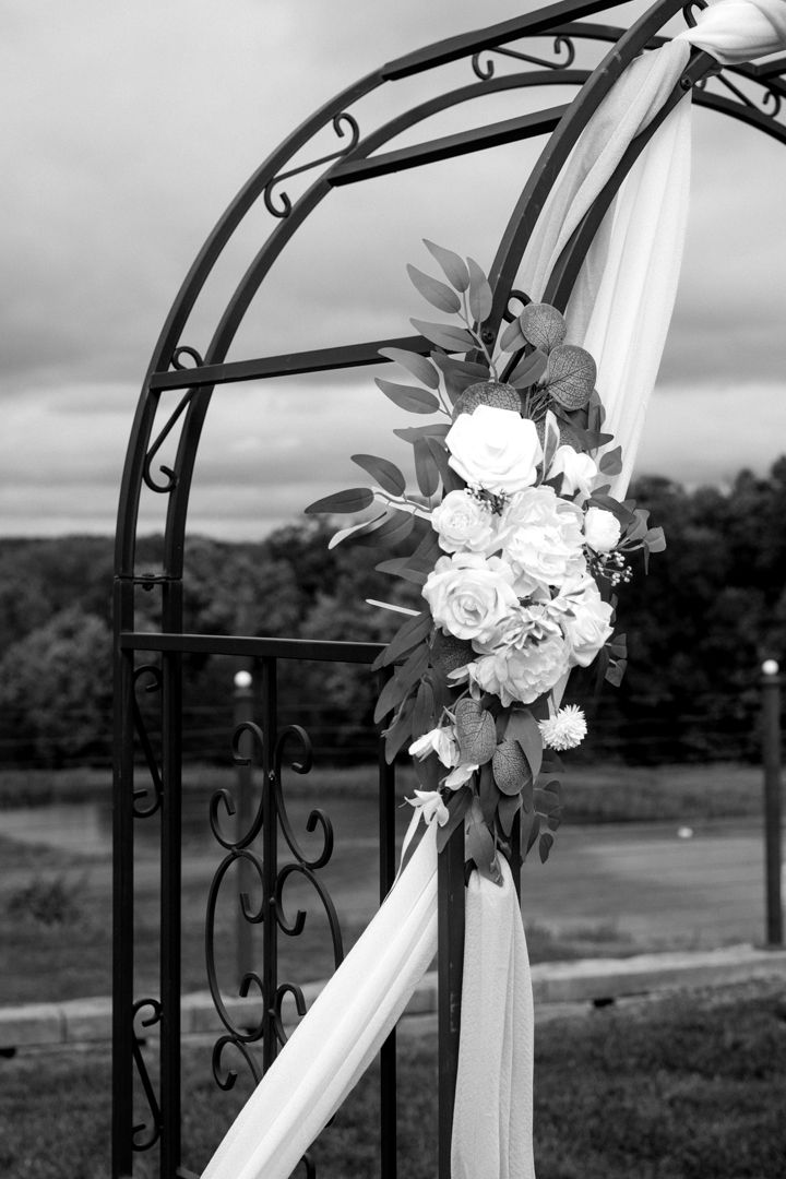 A black and white photo of a metal wedding arch decorated with roses, greenery and draped fabric overlooking a lake and trees.