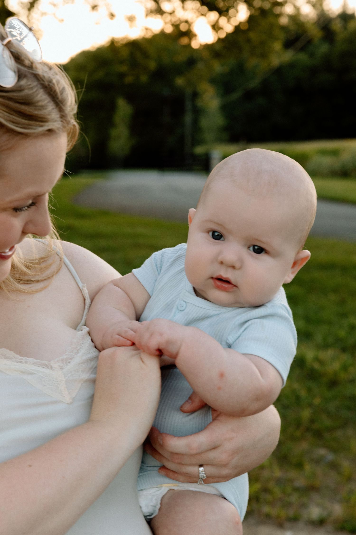 Smiling mother in a white lace camisole holds a chubby baby in a light blue outfit outdoors on a grassy lawn at golden hour.