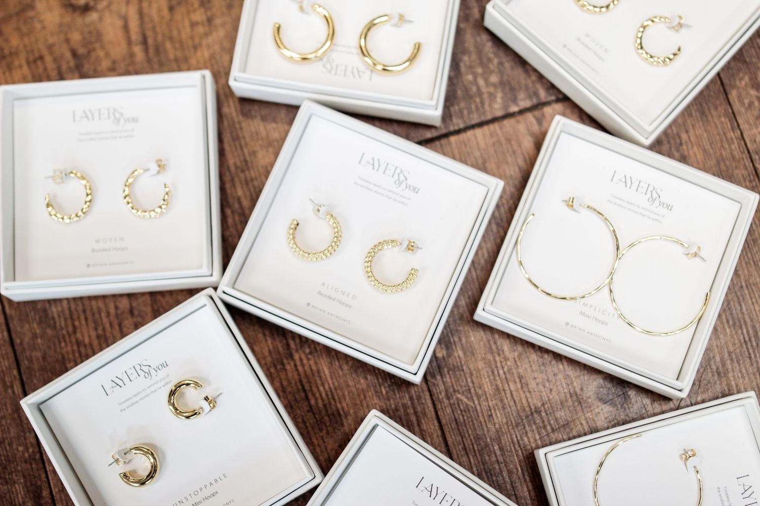 Gold hoop earrings in white gift boxes arranged on a rustic wooden table, featuring thick, twisted, and thin hoop styles.