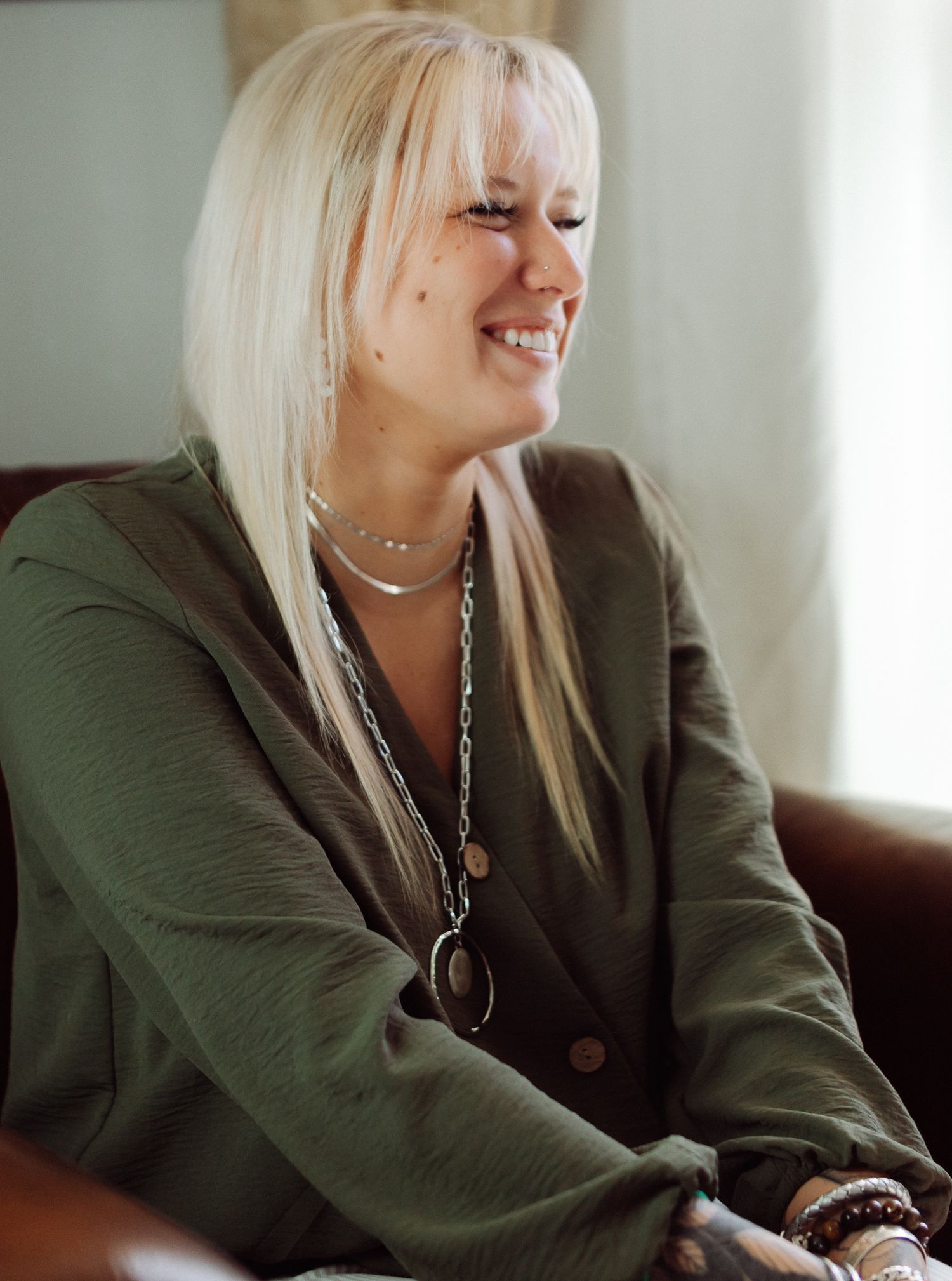 Smiling woman with long platinum blonde hair wears an olive green cardigan and layered necklaces while seated on a leather couch.