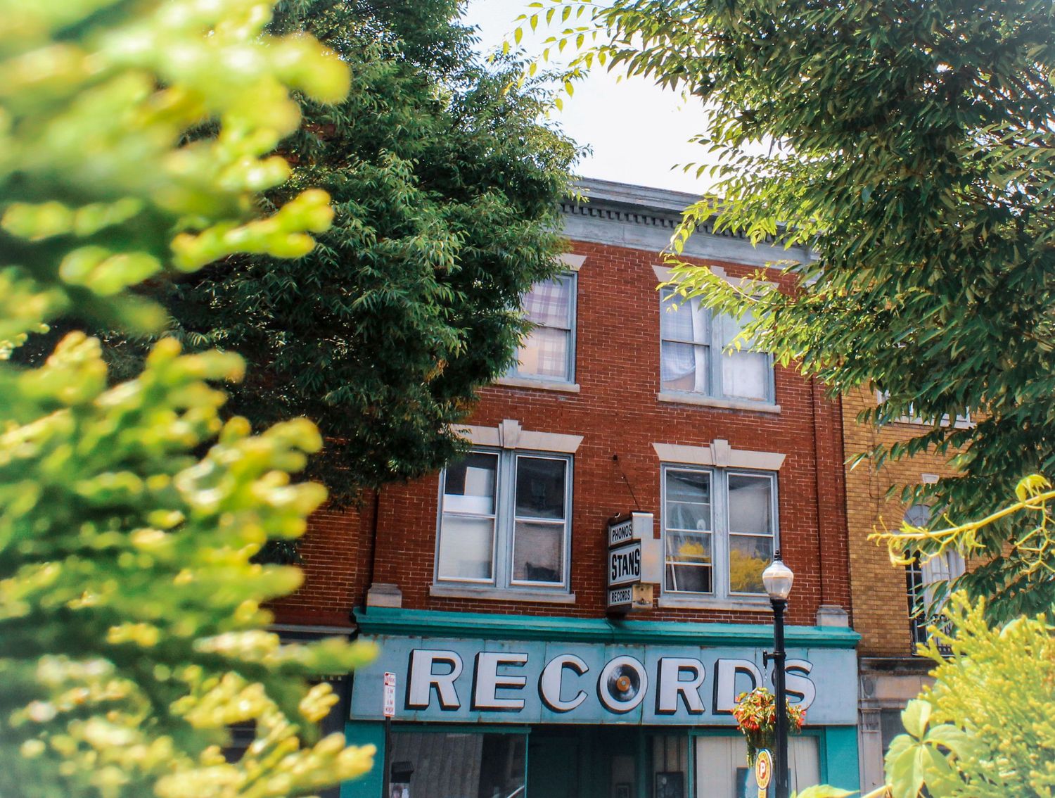 Red brick building with a teal storefront and large RECORDS sign, framed by lush green summer tree leaves on a sunny day.