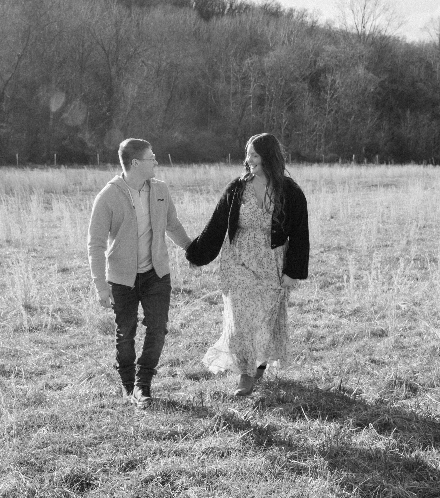 Black and white photo of a smiling couple holding hands while walking through a grassy open field with trees in the background.