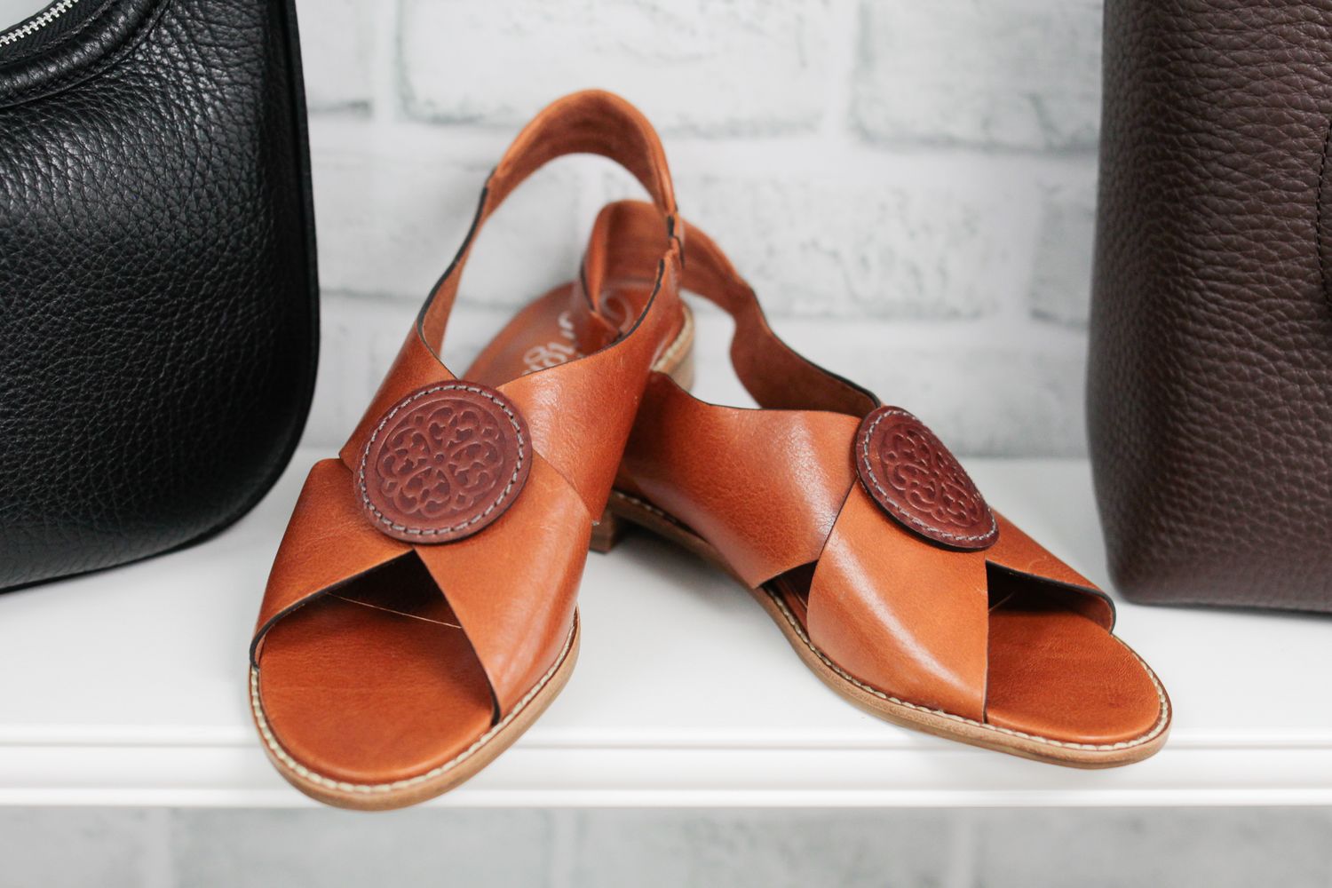 Pair of tan leather slingback sandals with crisscross straps and round medallion details displayed on a white shelf near handbags.