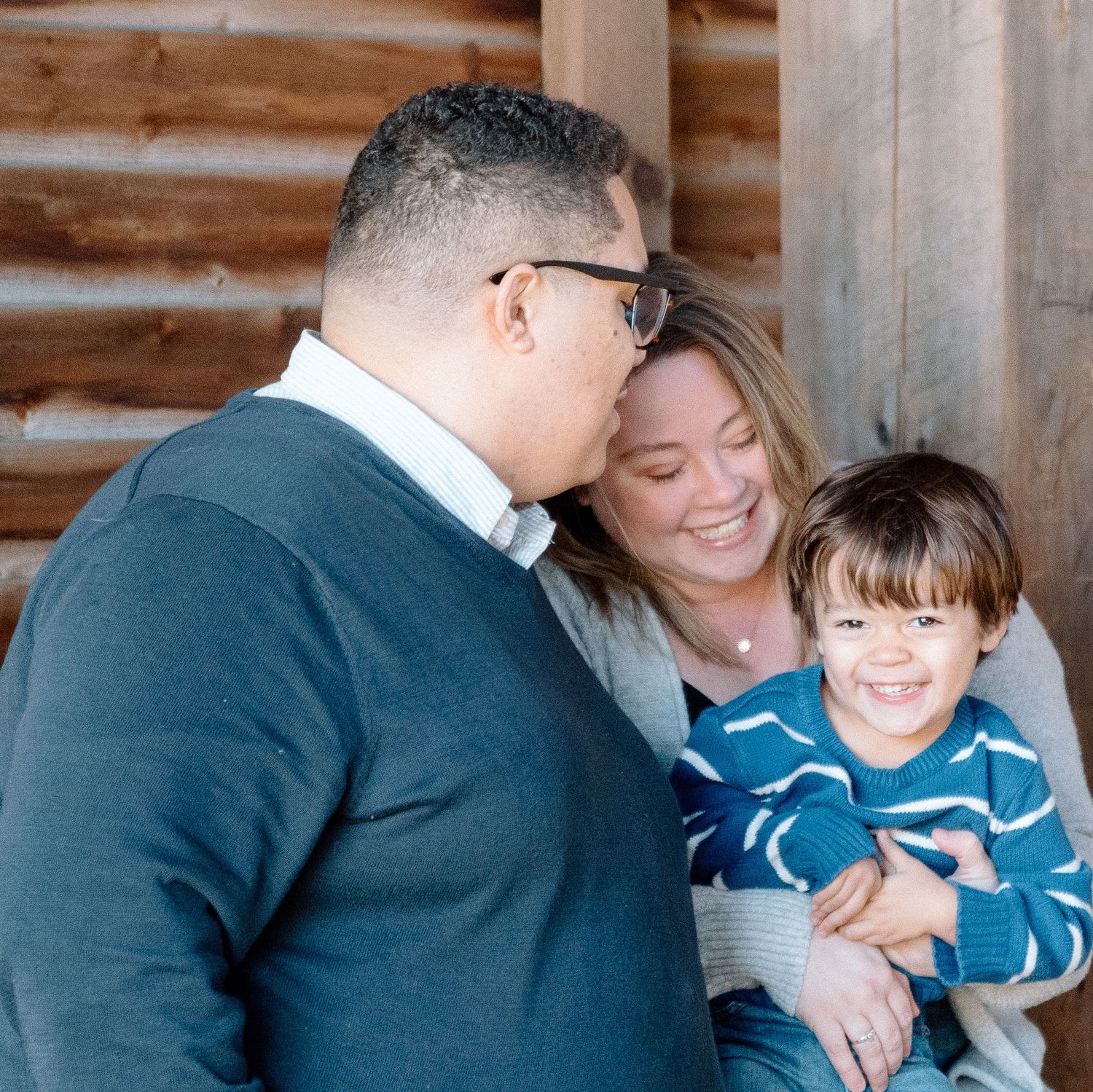A smiling family of three poses against a rustic log cabin wall as a man kisses a woman's cheek while she holds a laughing toddler.