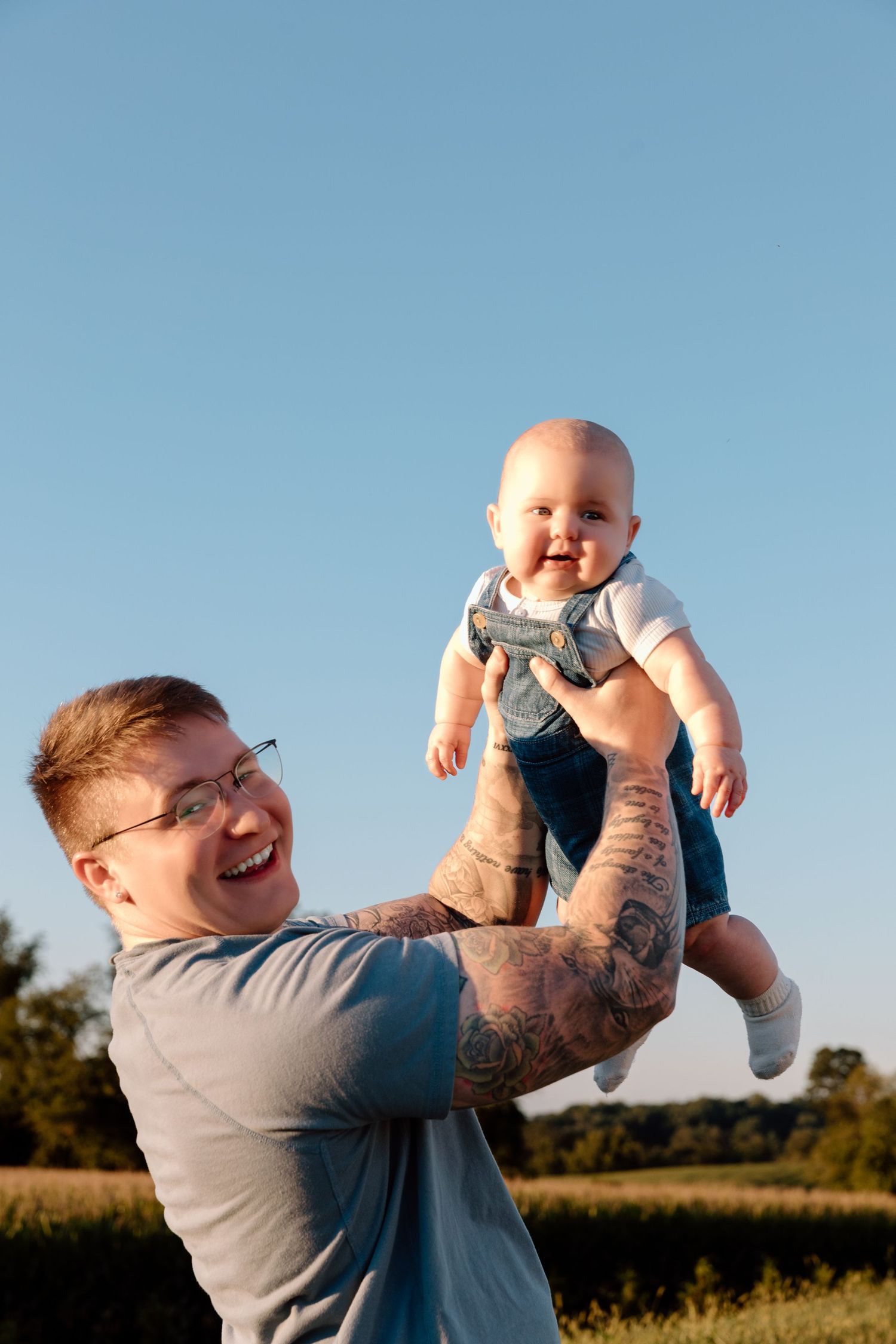 Smiling tattooed man with glasses lifts a laughing baby in denim overalls into the air against a clear blue sky in a sunny field.